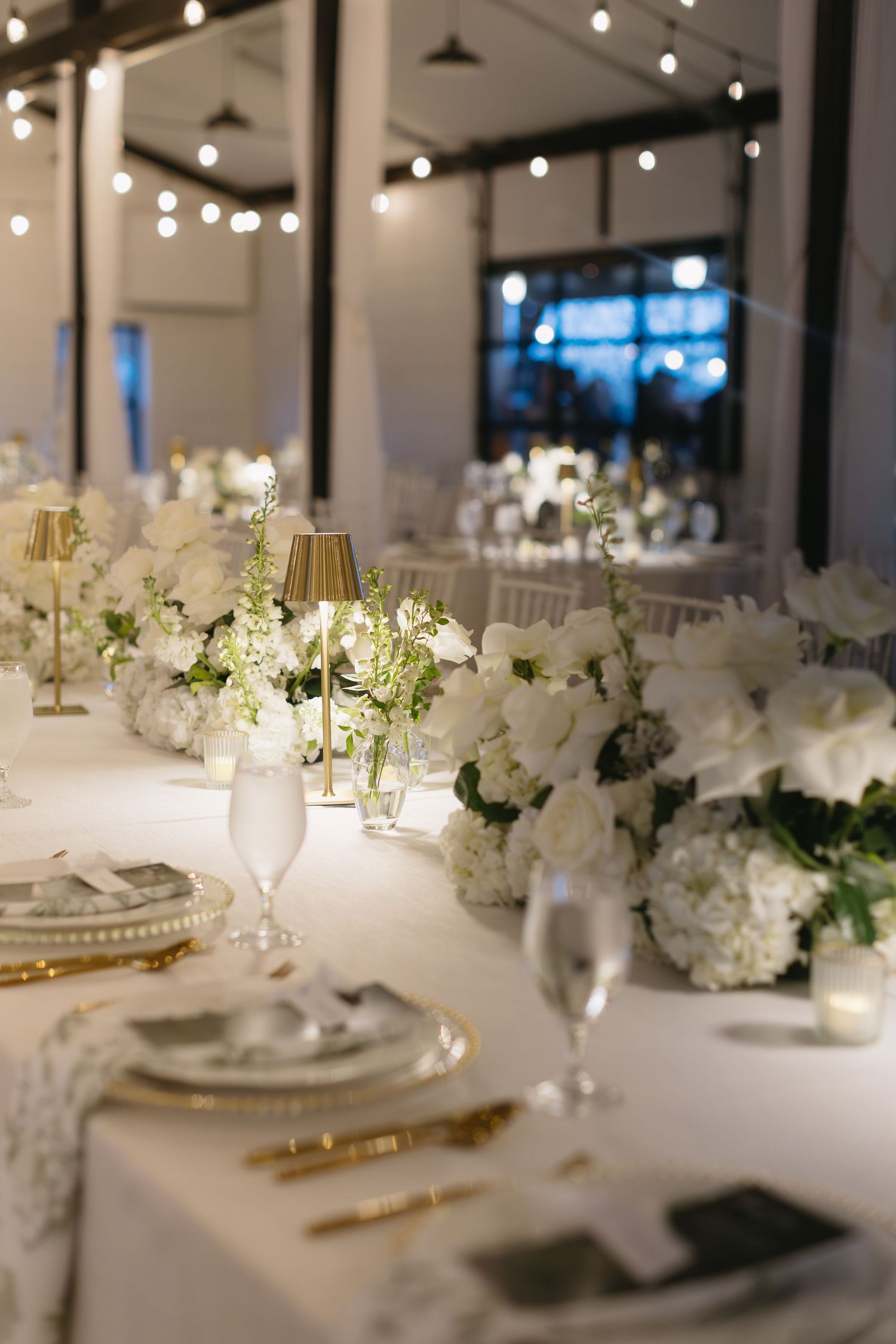 Long wedding reception table with white flowers, gold accents, and string lights creating a romantic ambiance.