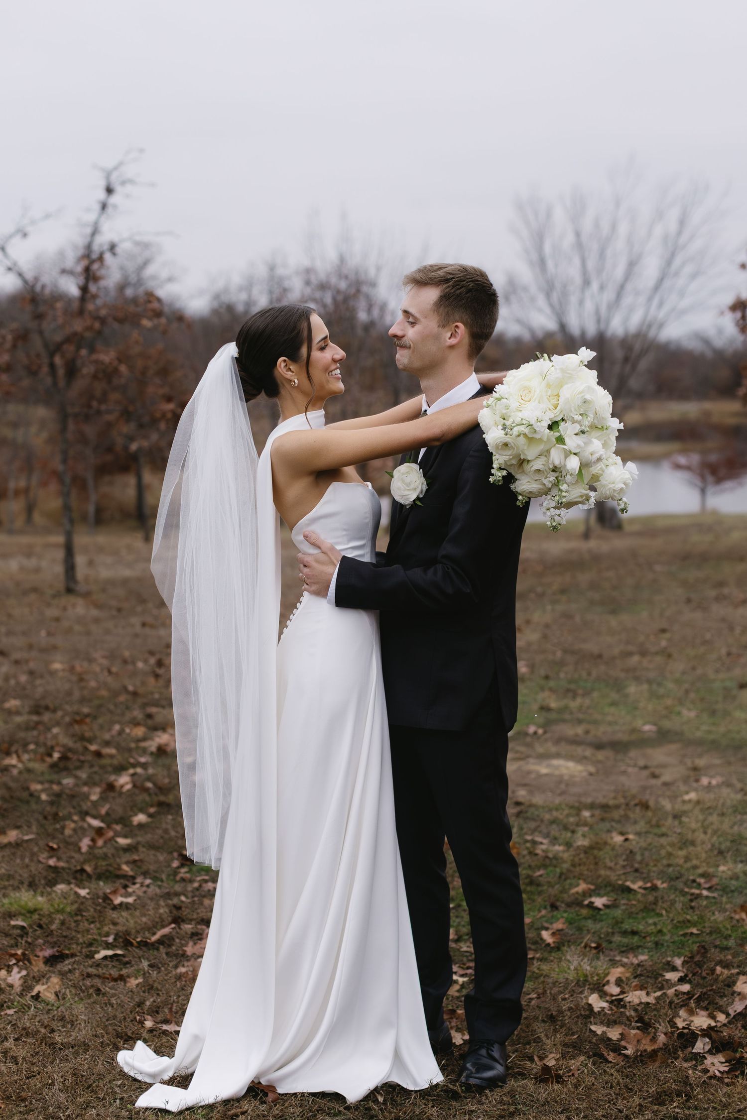 A bride in a white gown holds a bouquet of white flowers while embracing in an outdoor setting.