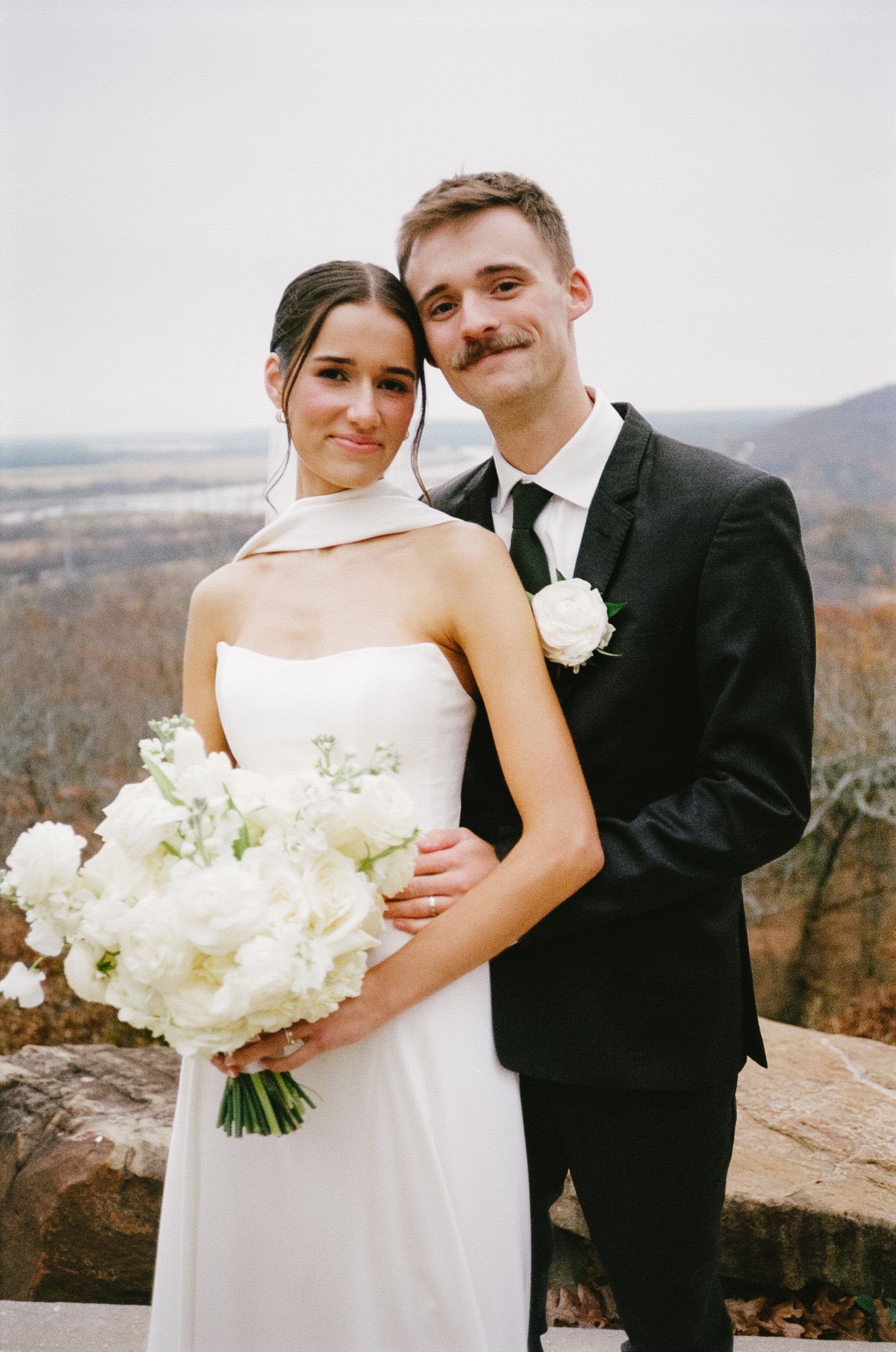 A couple in wedding attire pose with white flowers against a scenic mountain backdrop.