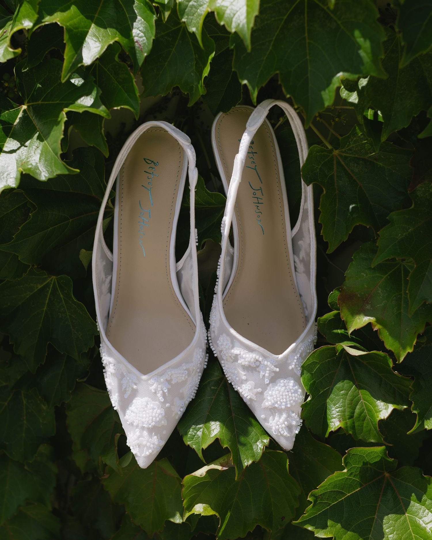 Delicate white bridal shoes with lace details arranged against a backdrop of lush green ivy leaves.