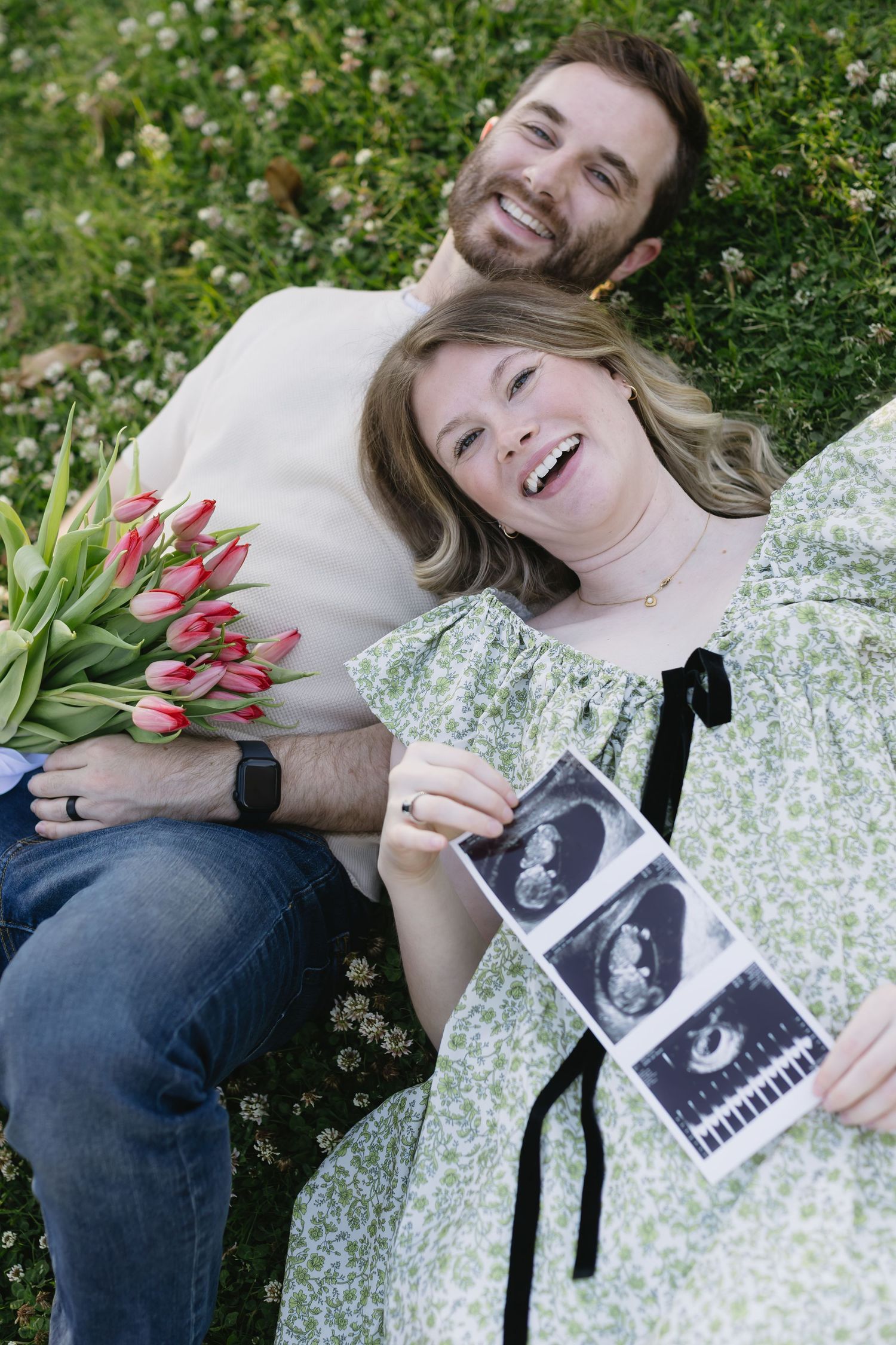 Couple lying on grass sharing joyful moments while holding ultrasound photos in a garden.