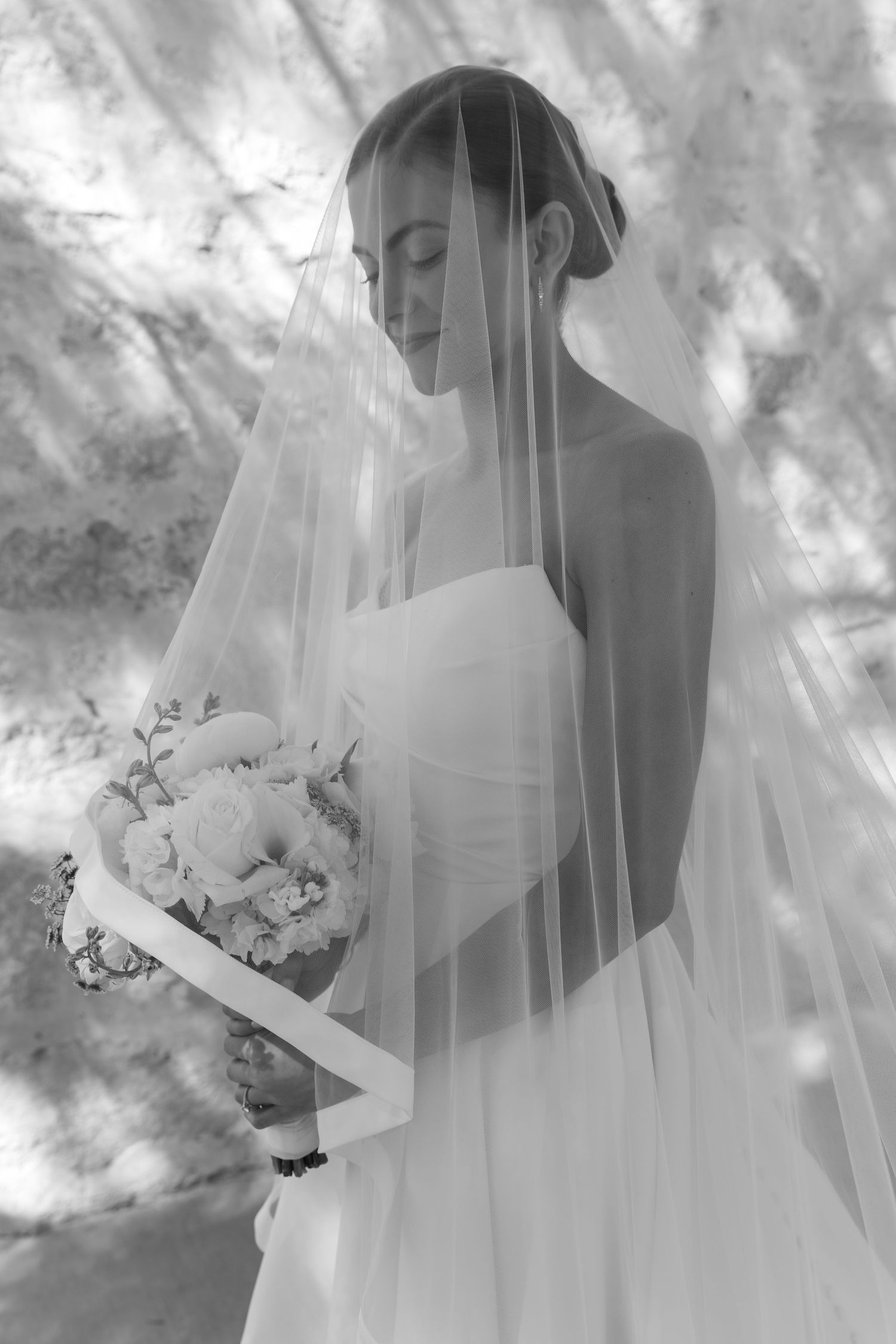 Ethereal black and white portrait of a bride in a strapless gown holding a bouquet under a flowing veil.