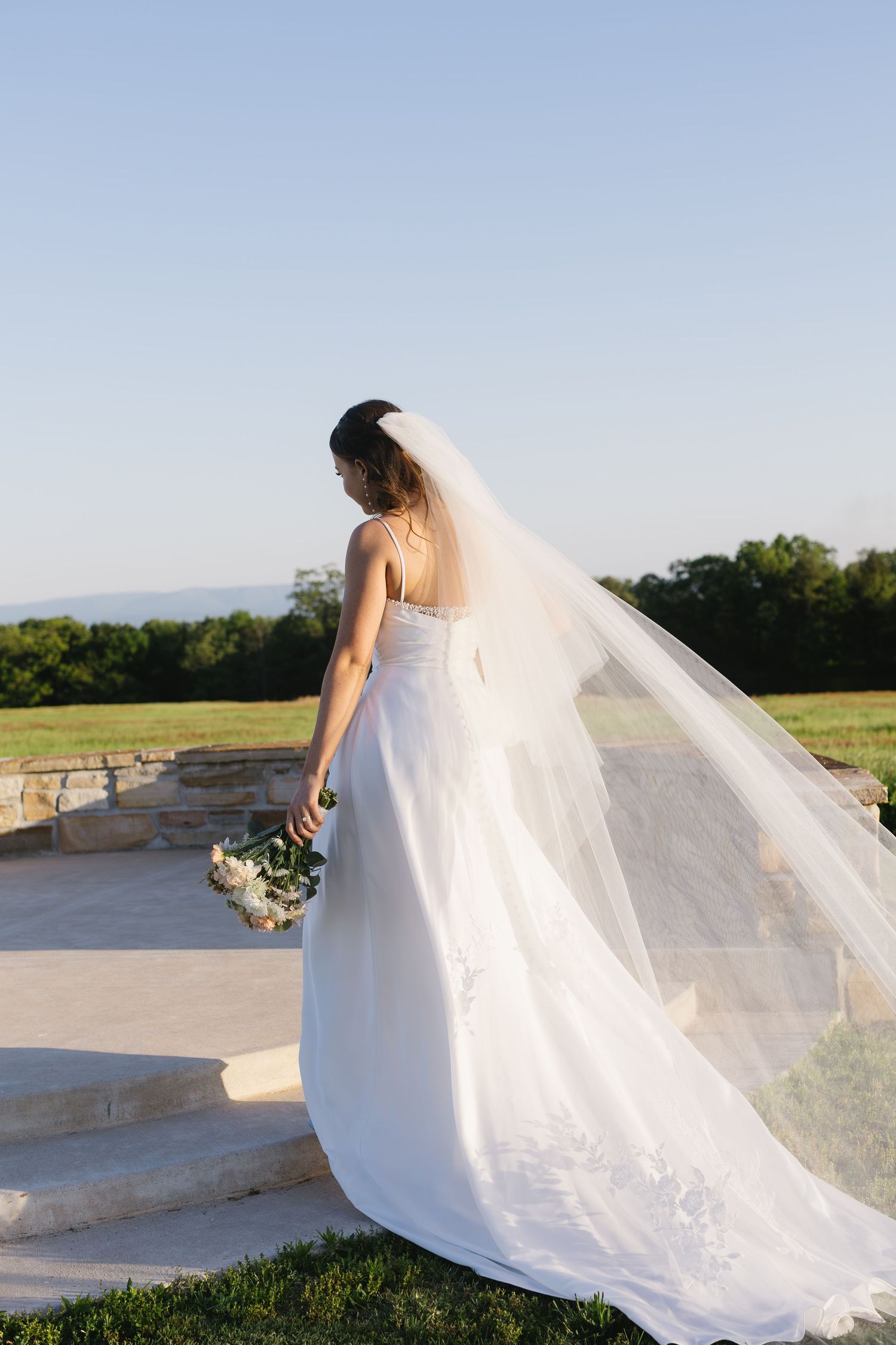 A bride in a flowing white gown stands gracefully on stone steps with her veil catching the breeze.