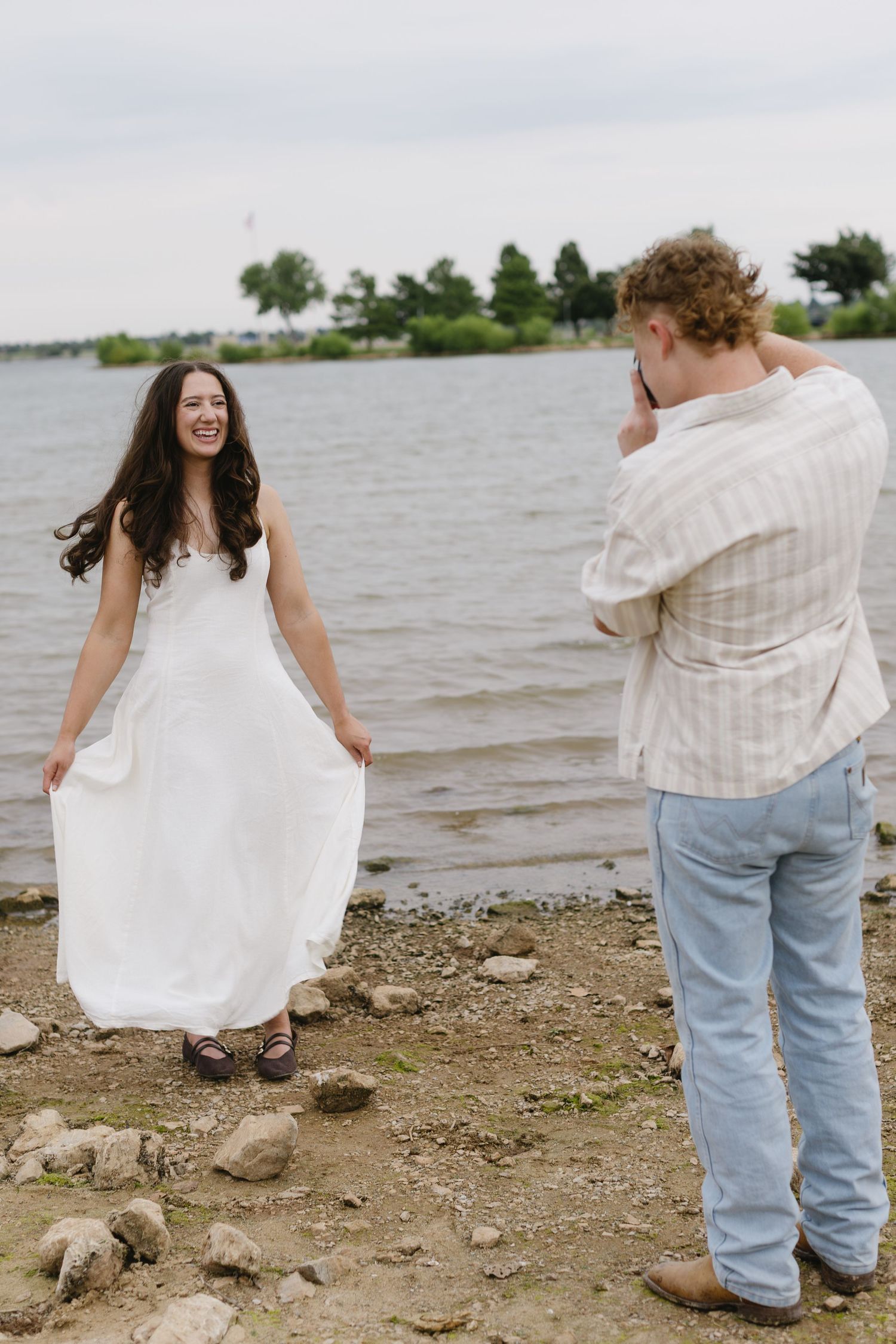 Two people in casual clothing throw rocks into a lake from a pebbly shoreline.