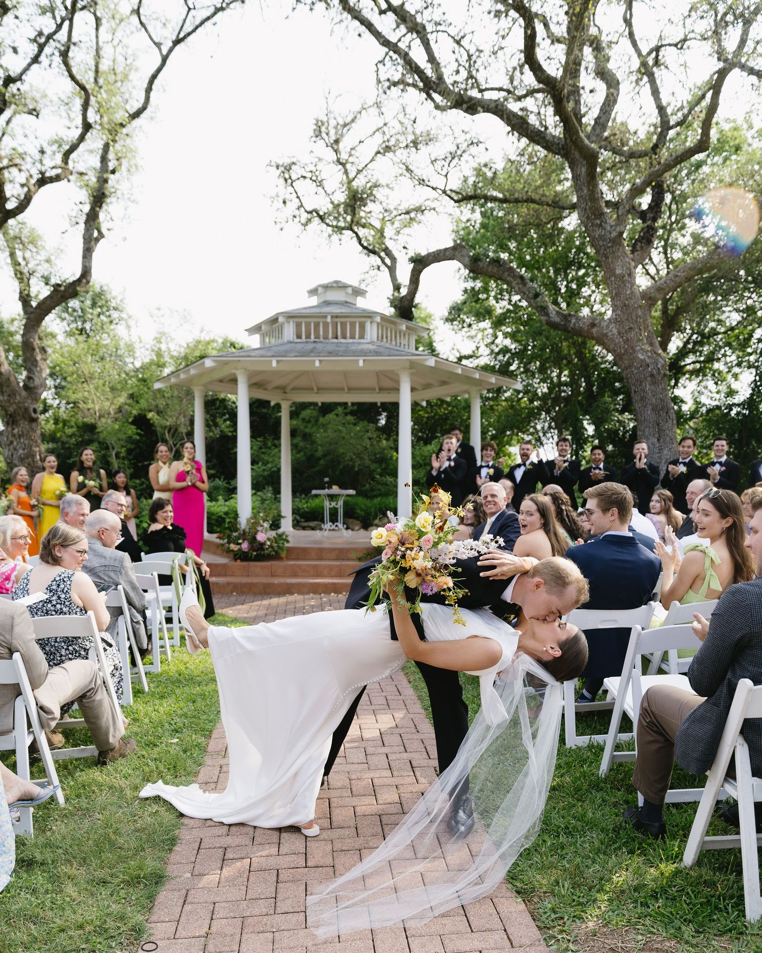 Outdoor wedding ceremony setup with white chairs and floral arrangements under large oak trees.