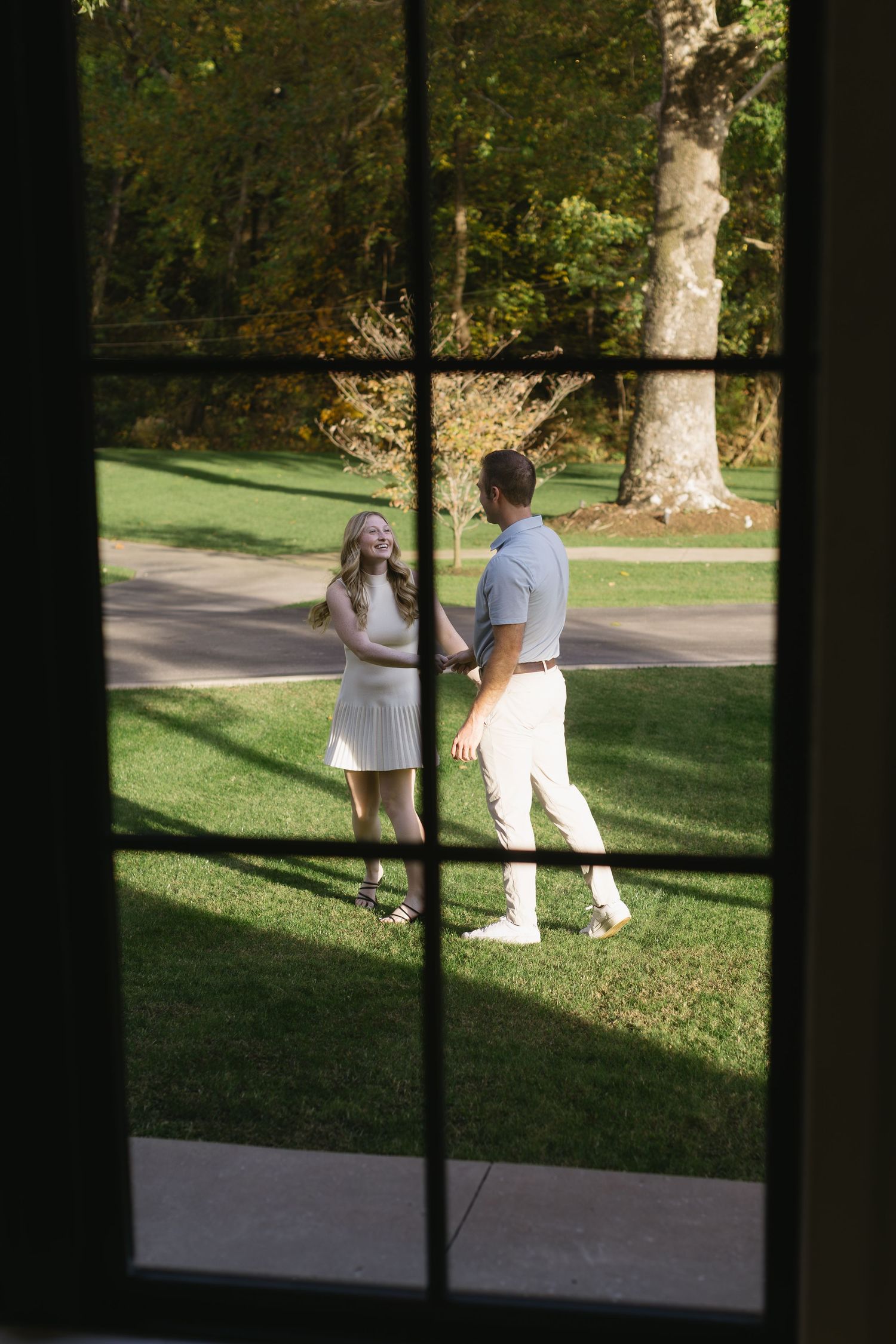 A romantic scene viewed through a window pane shows a couple standing together on a sunlit lawn.