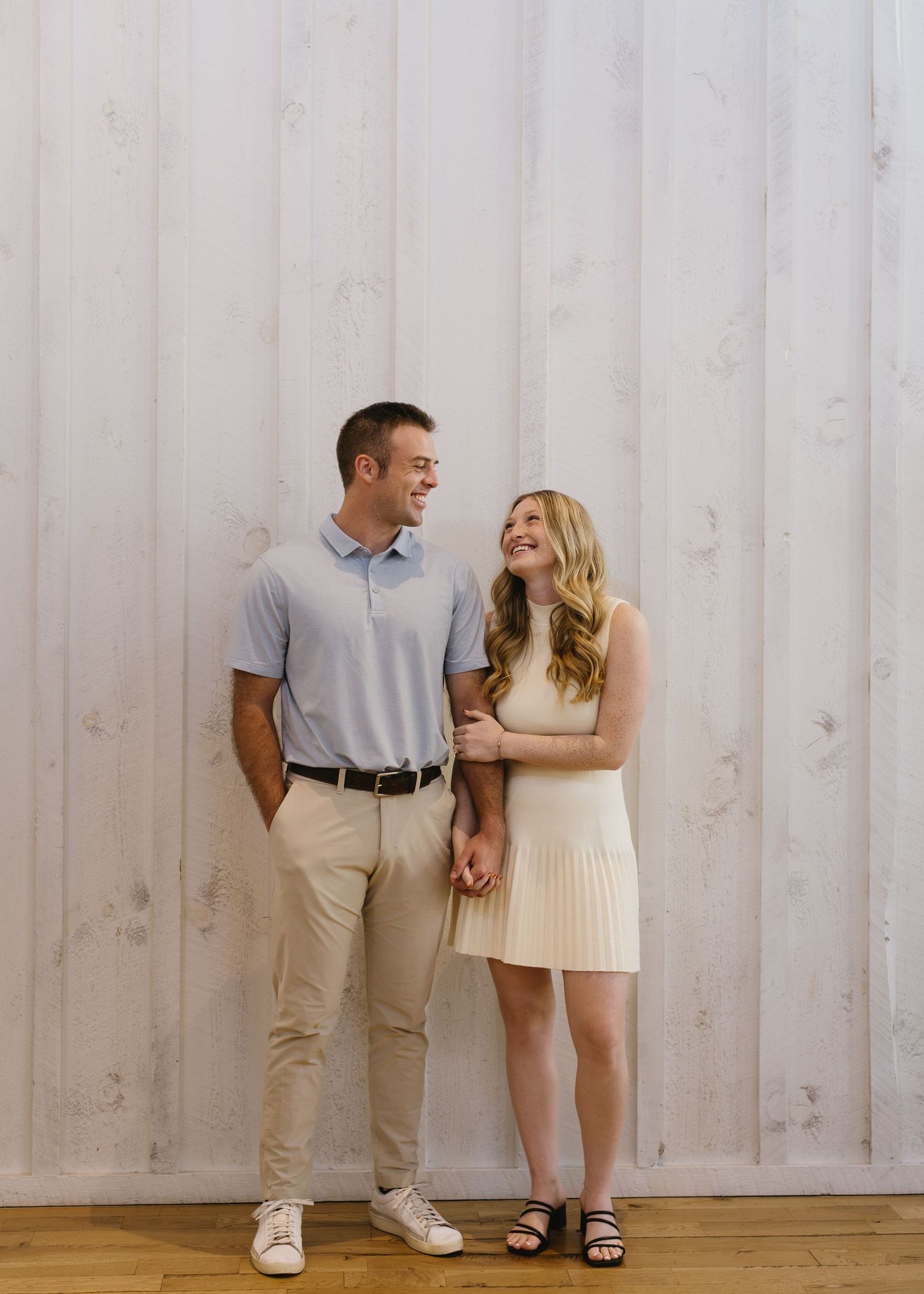 Couple in casual summer outfits poses together against a white wall background.