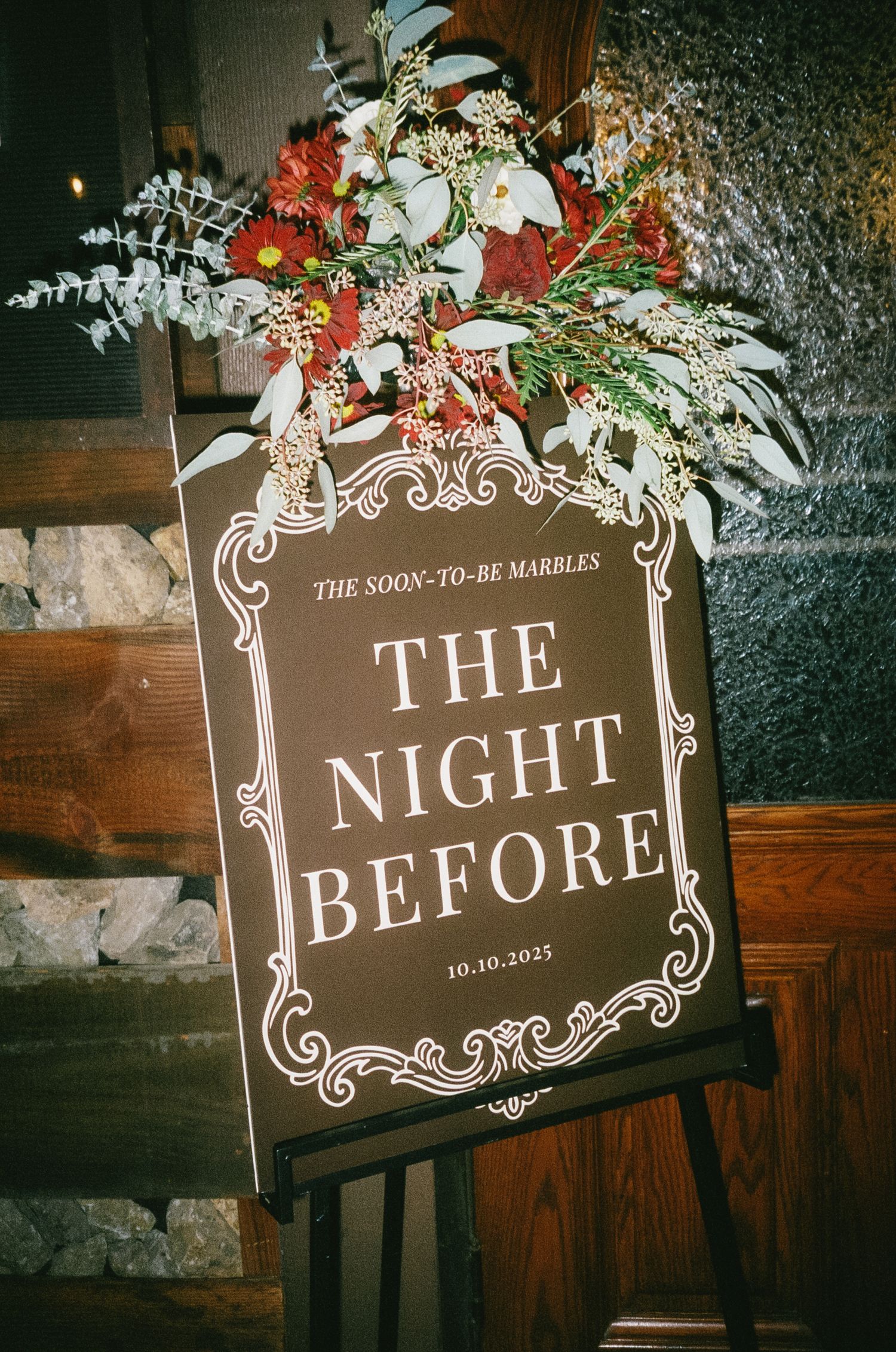 A wedding rehearsal dinner sign reading 'The Night Before' decorated with red and white floral arrangements.