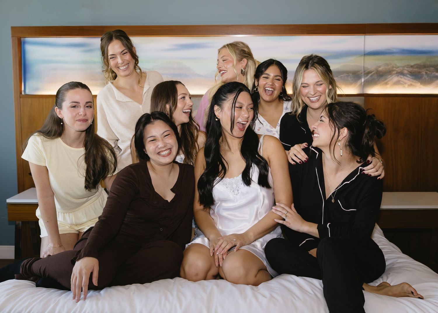 A group of friends in black and white outfits laugh together while lounging on a bed during a wedding celebration.