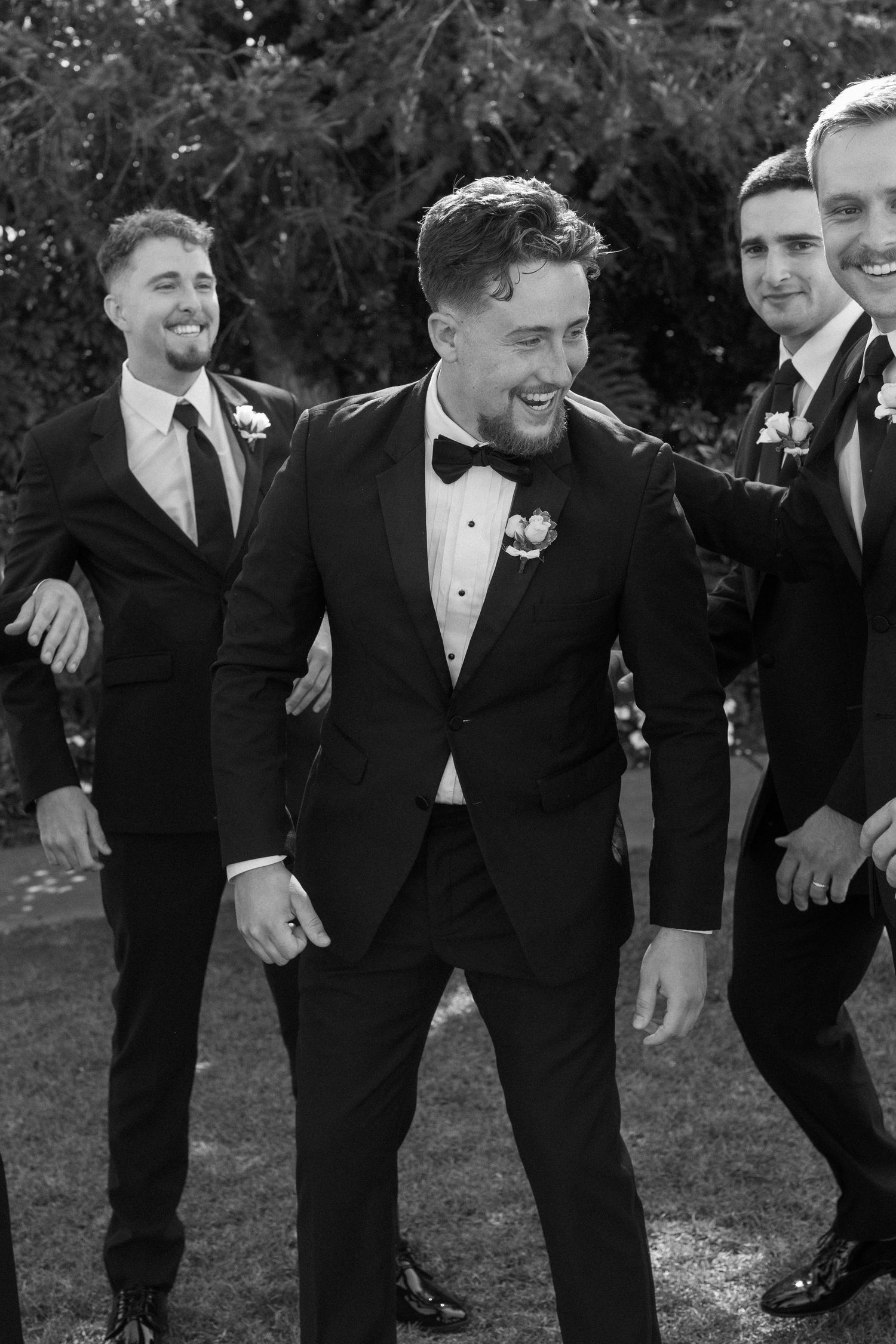 Groomsmen in black tuxedos share candid laughing moments during outdoor wedding photography.