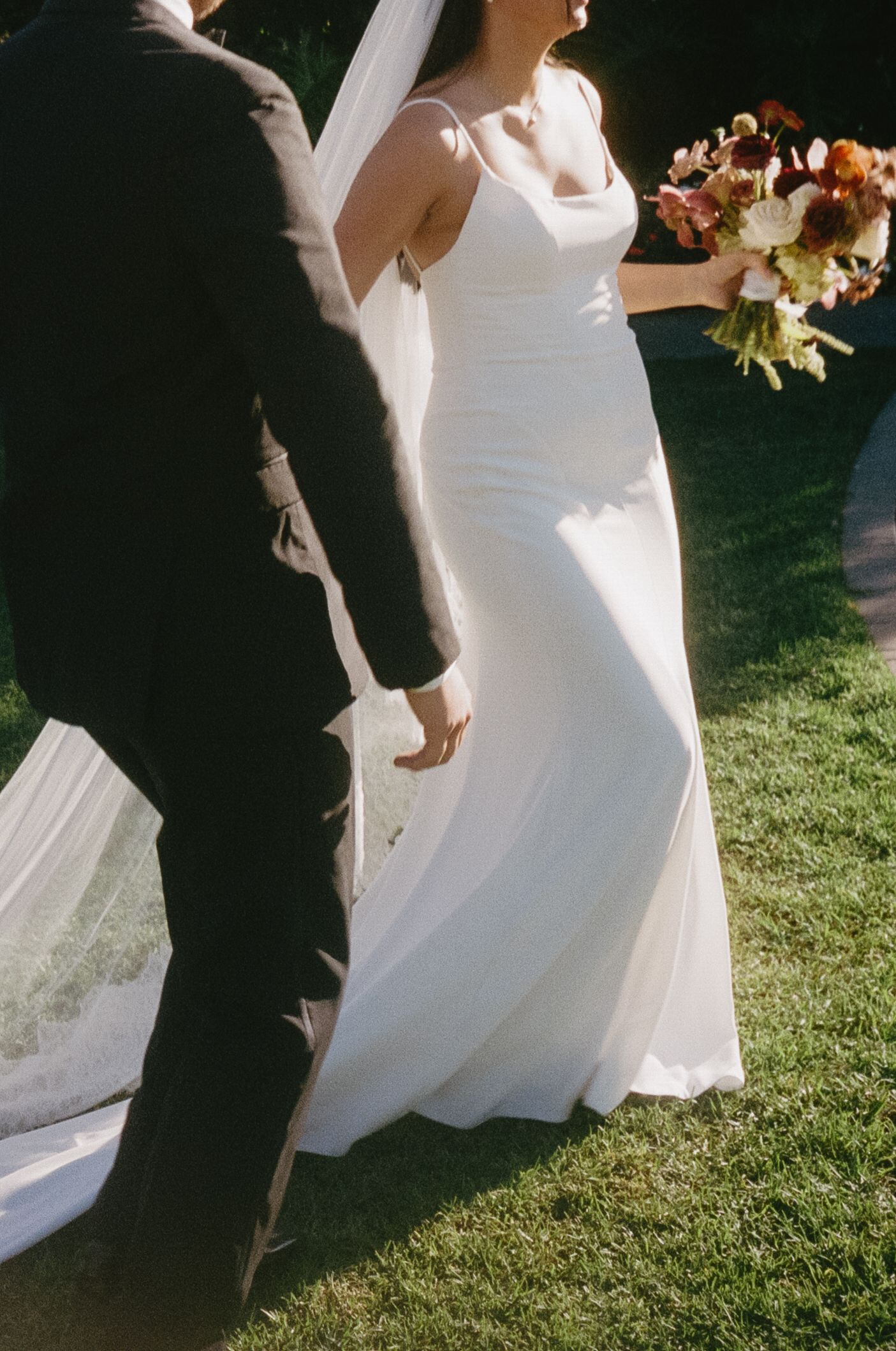 A wedding couple poses on green grass in elegant attire with flowing white dress.