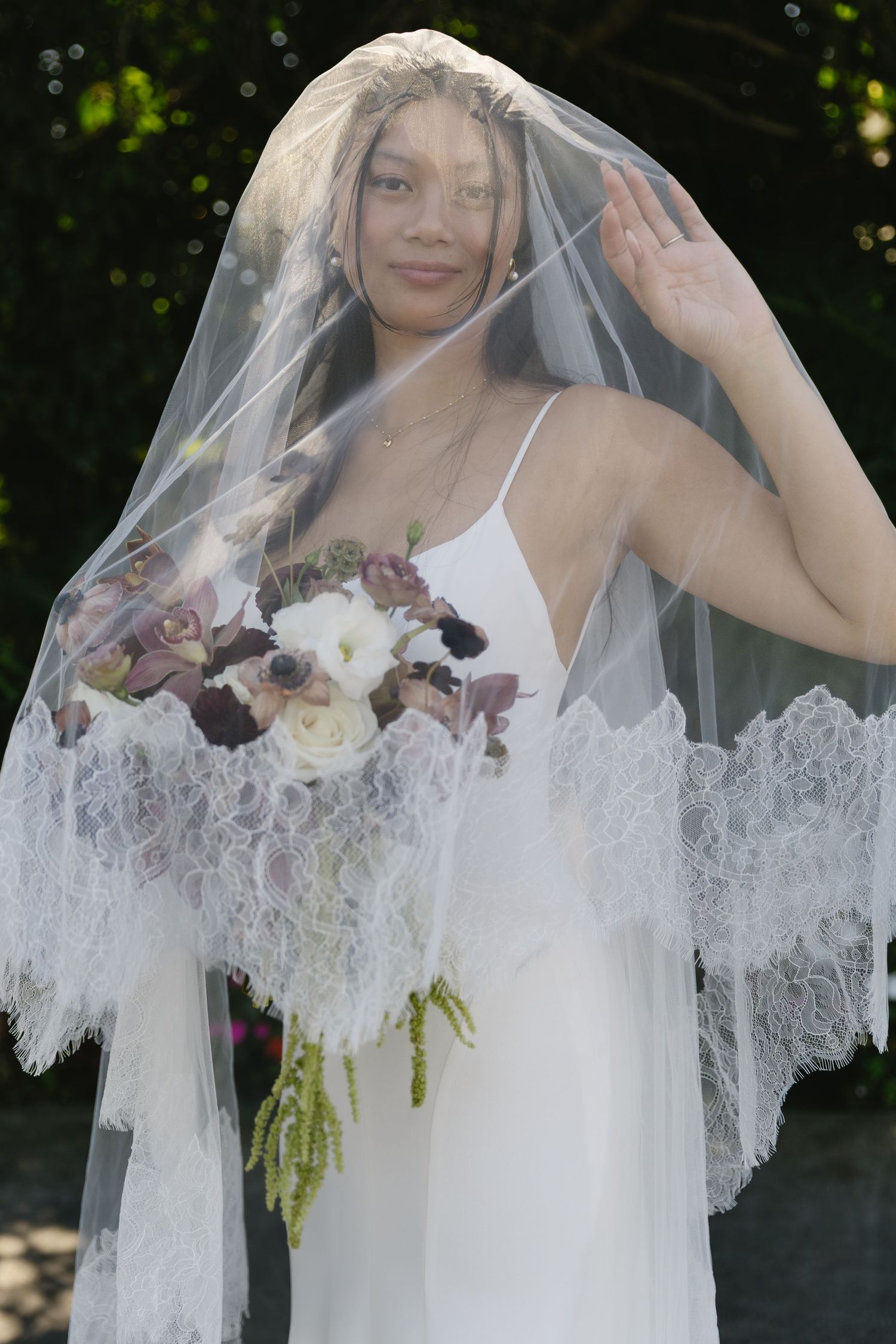 Bride with lace veil poses with elegant bouquet featuring white and purple blooms.
