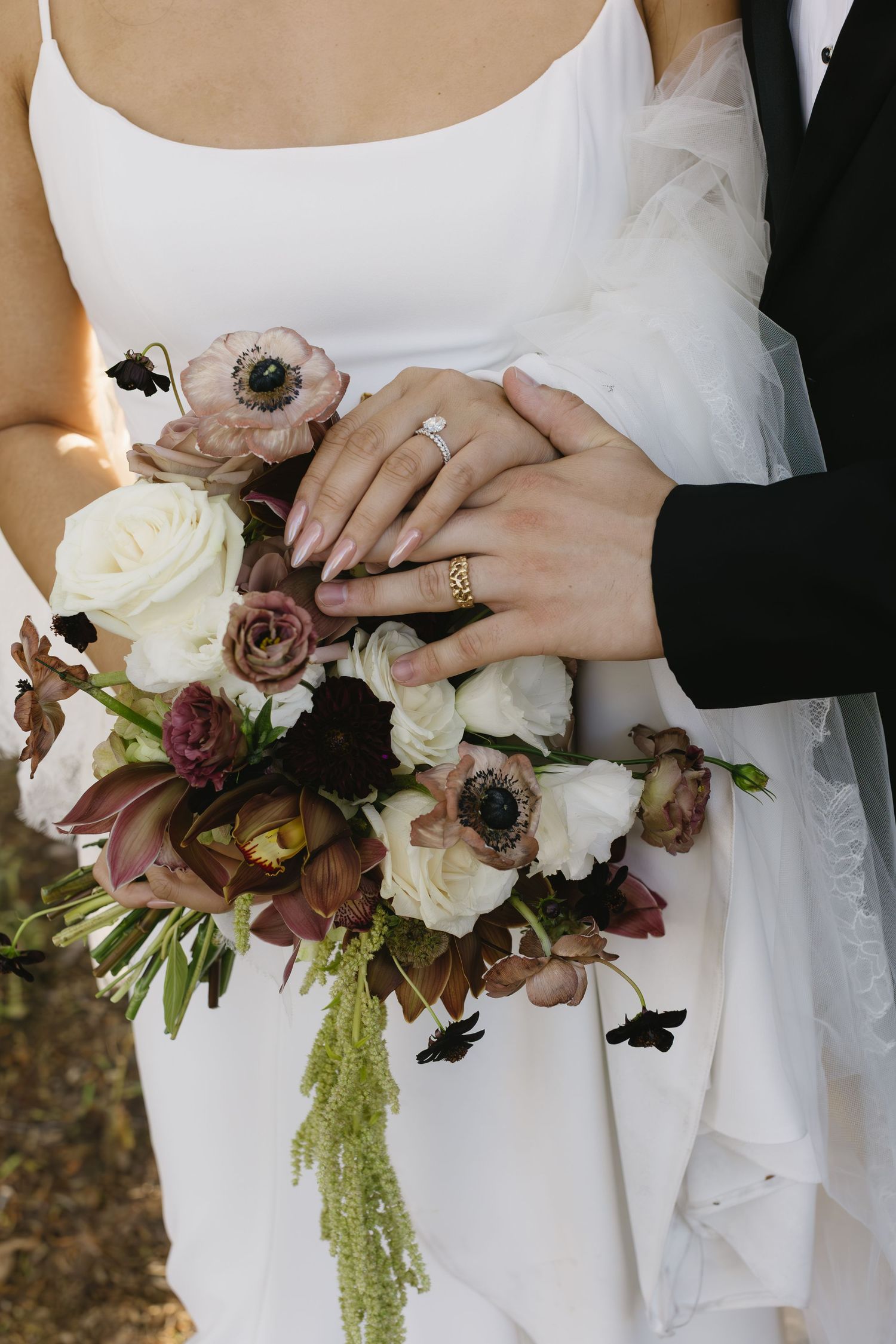 Close-up of wedding bouquet featuring white roses and burgundy flowers held by hands with rings.