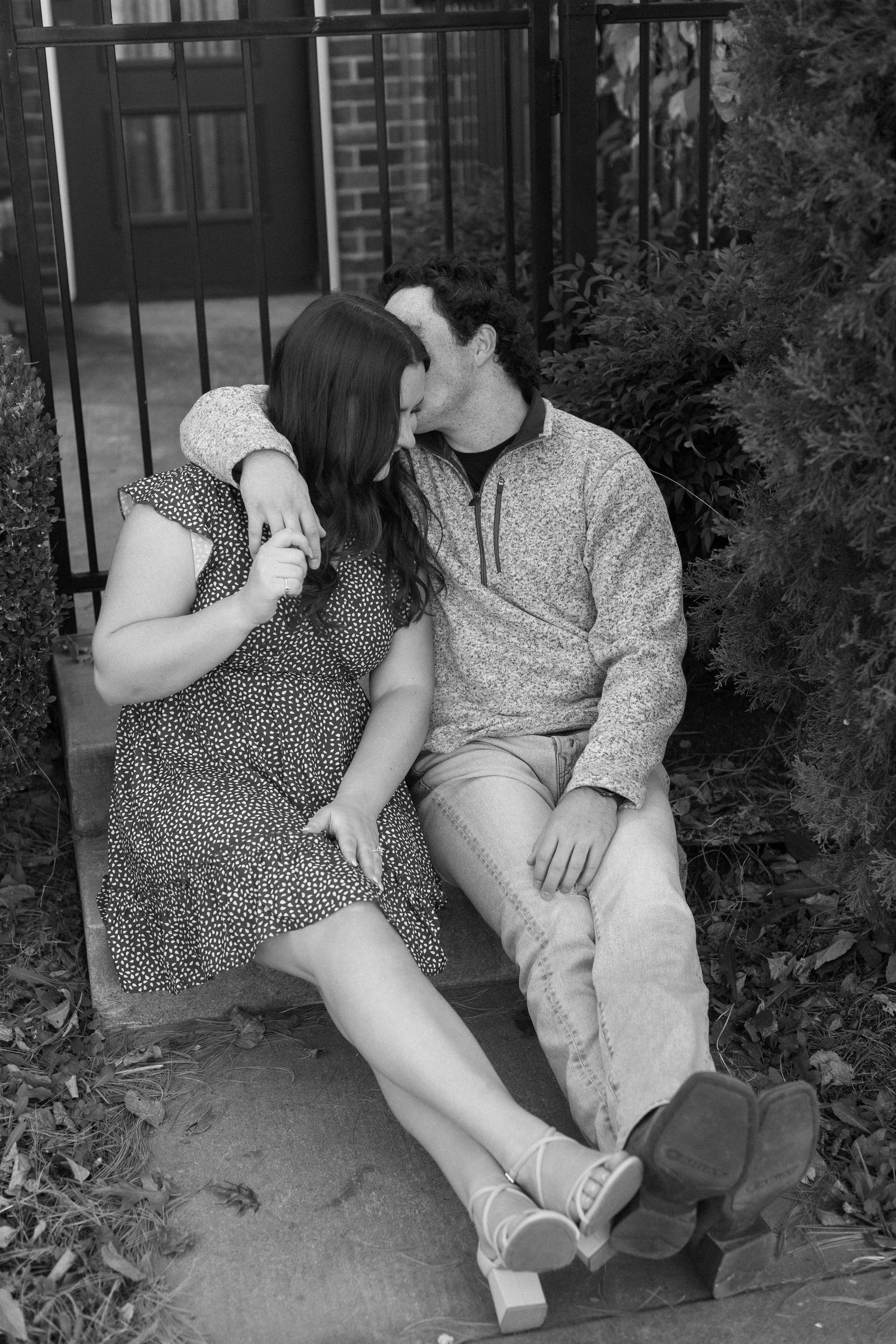 A black and white photo captures an intimate moment between a couple sitting by a gate.