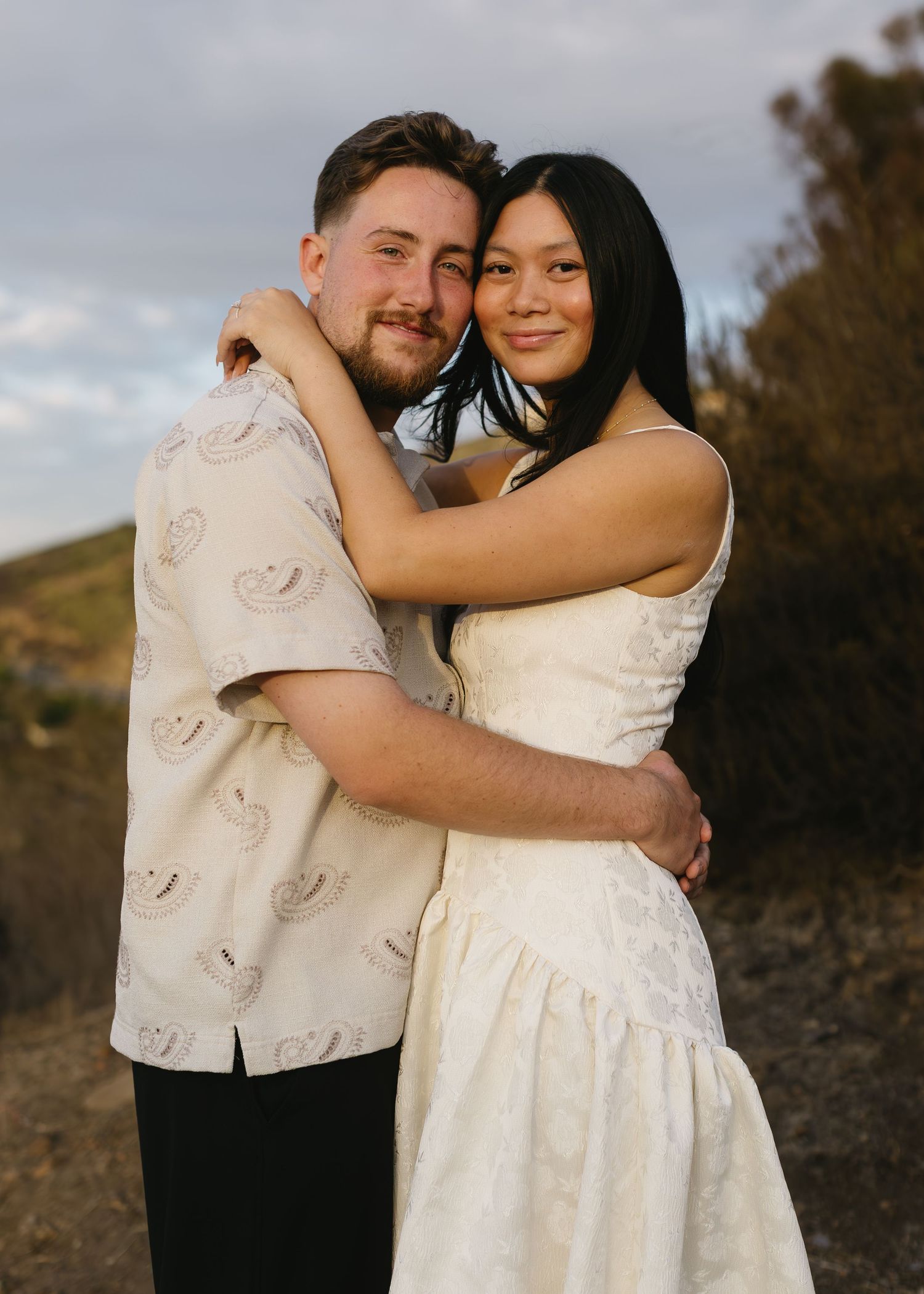 A couple embraces outdoors during a romantic photoshoot wearing white formal attire against a natural backdrop.