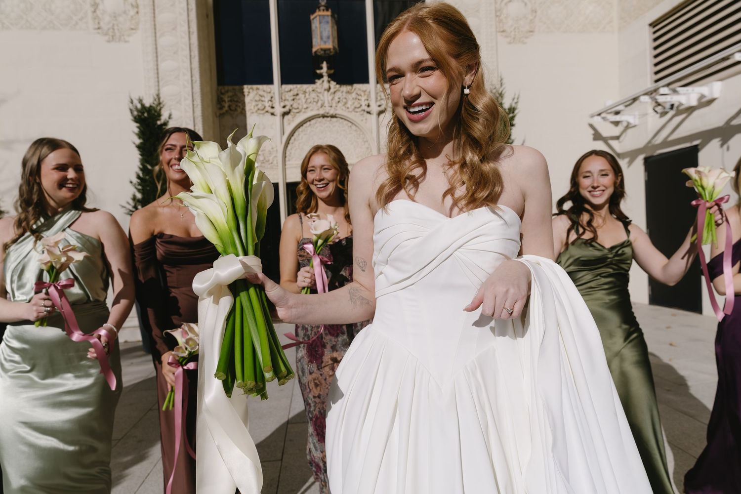 Joyful wedding party in mix-matched dresses celebrating with bouquets outdoors.