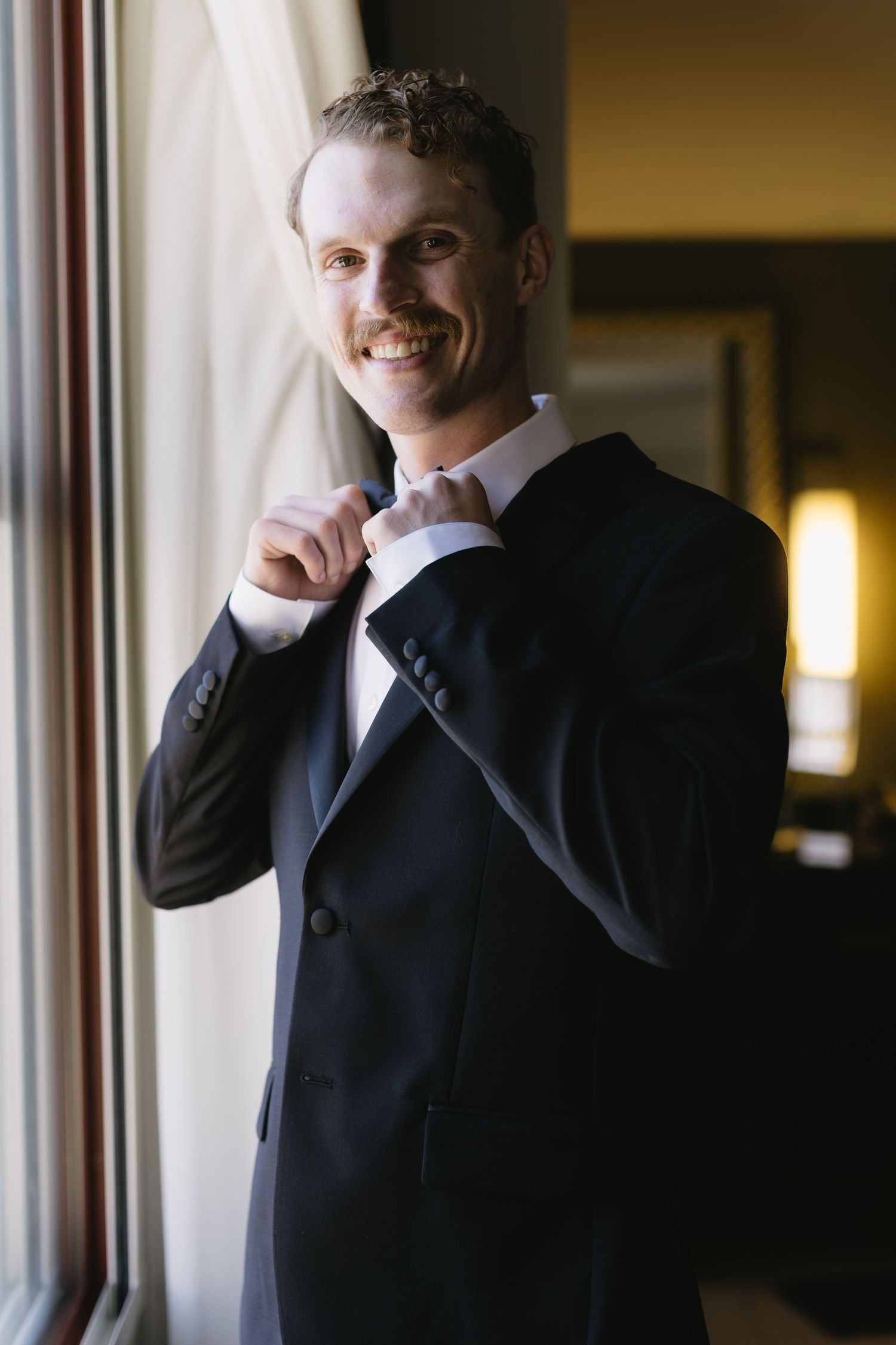 Person in dark suit adjusting tie near window with dramatic lighting.
