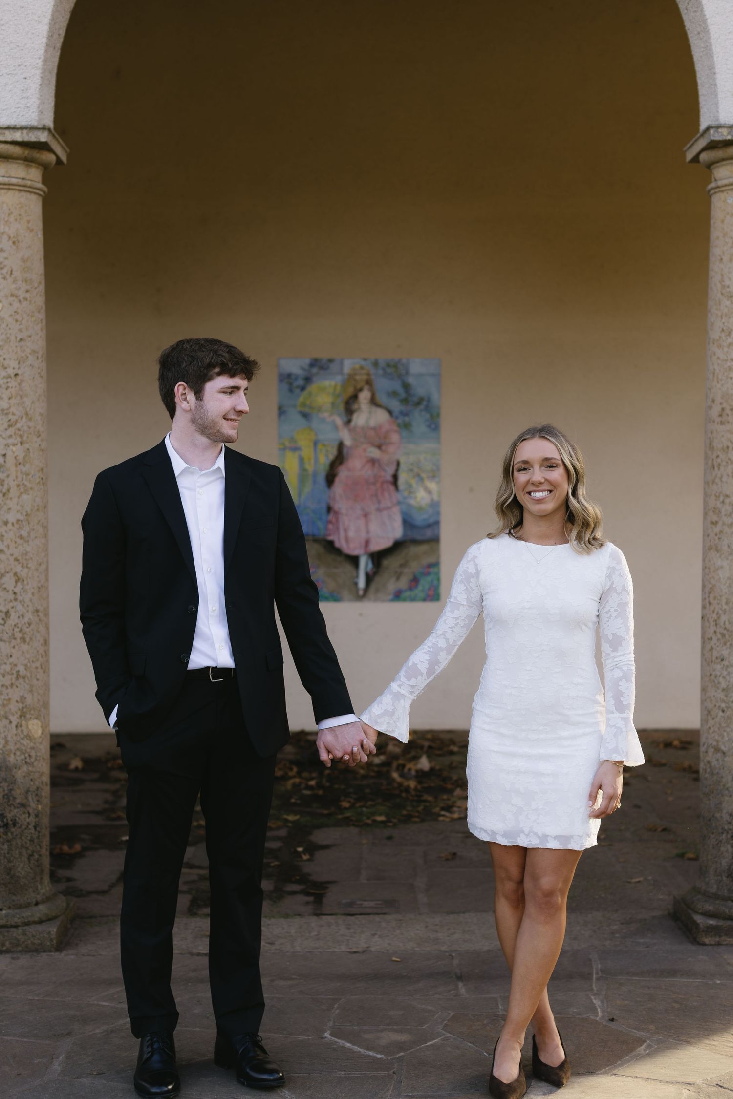 A couple in formal attire poses playfully in an outdoor colonnade setting.