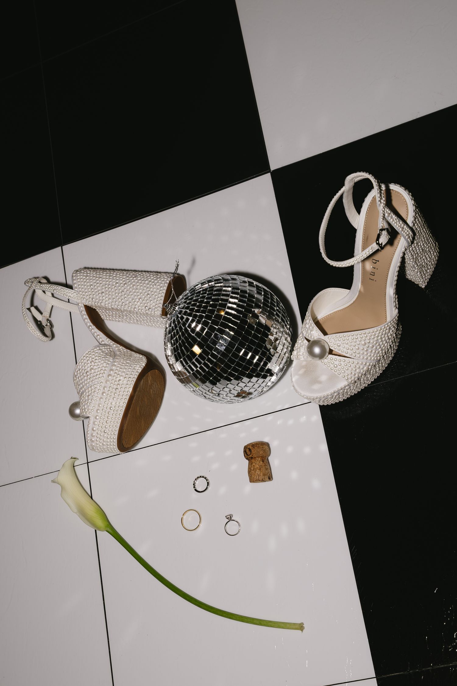 Wedding accessories including sparkly shoes, rings, and disco ball arranged artistically on white backdrop.