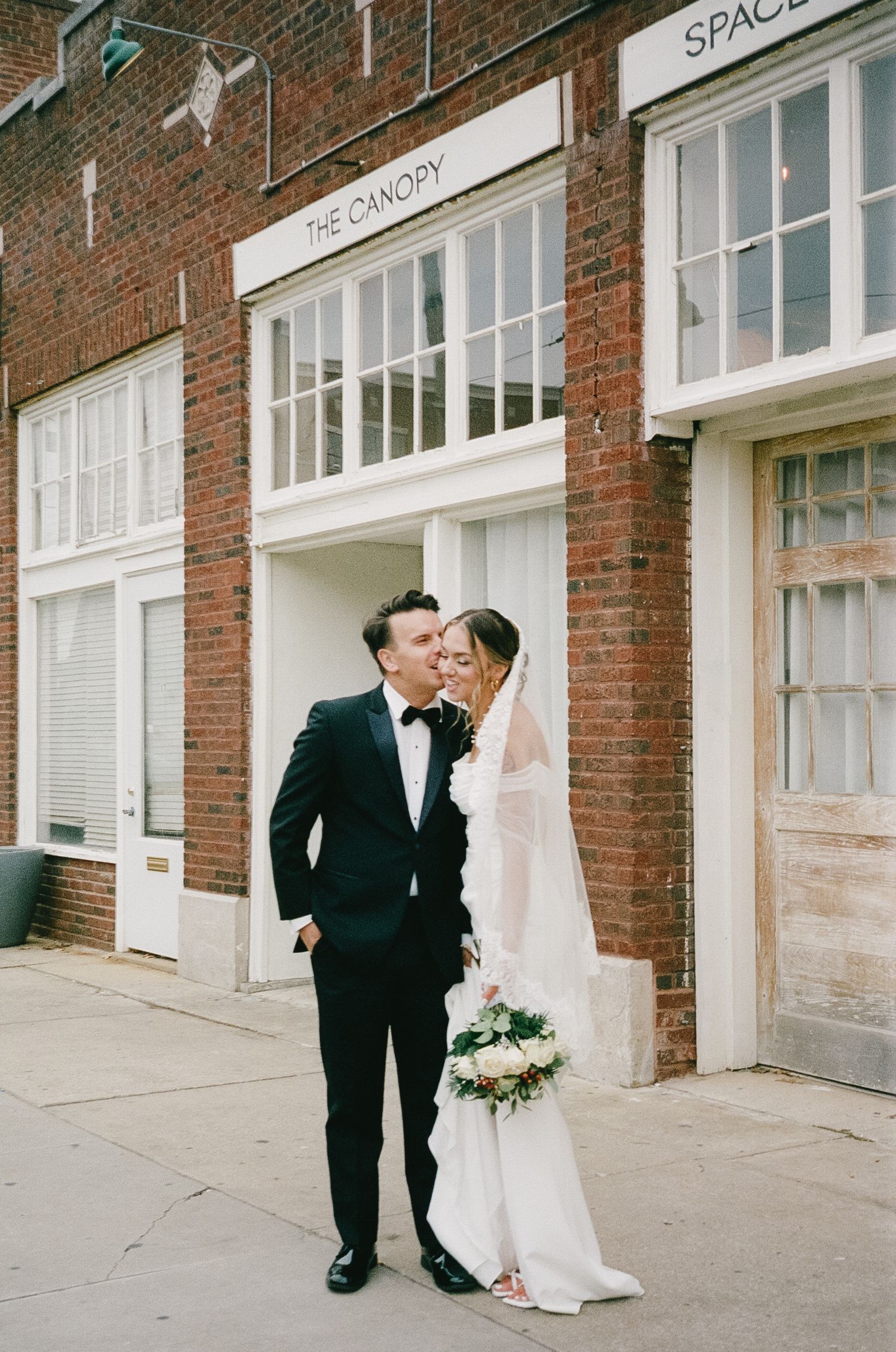 Wedding couple poses affectionately outside brick building with storefront windows.