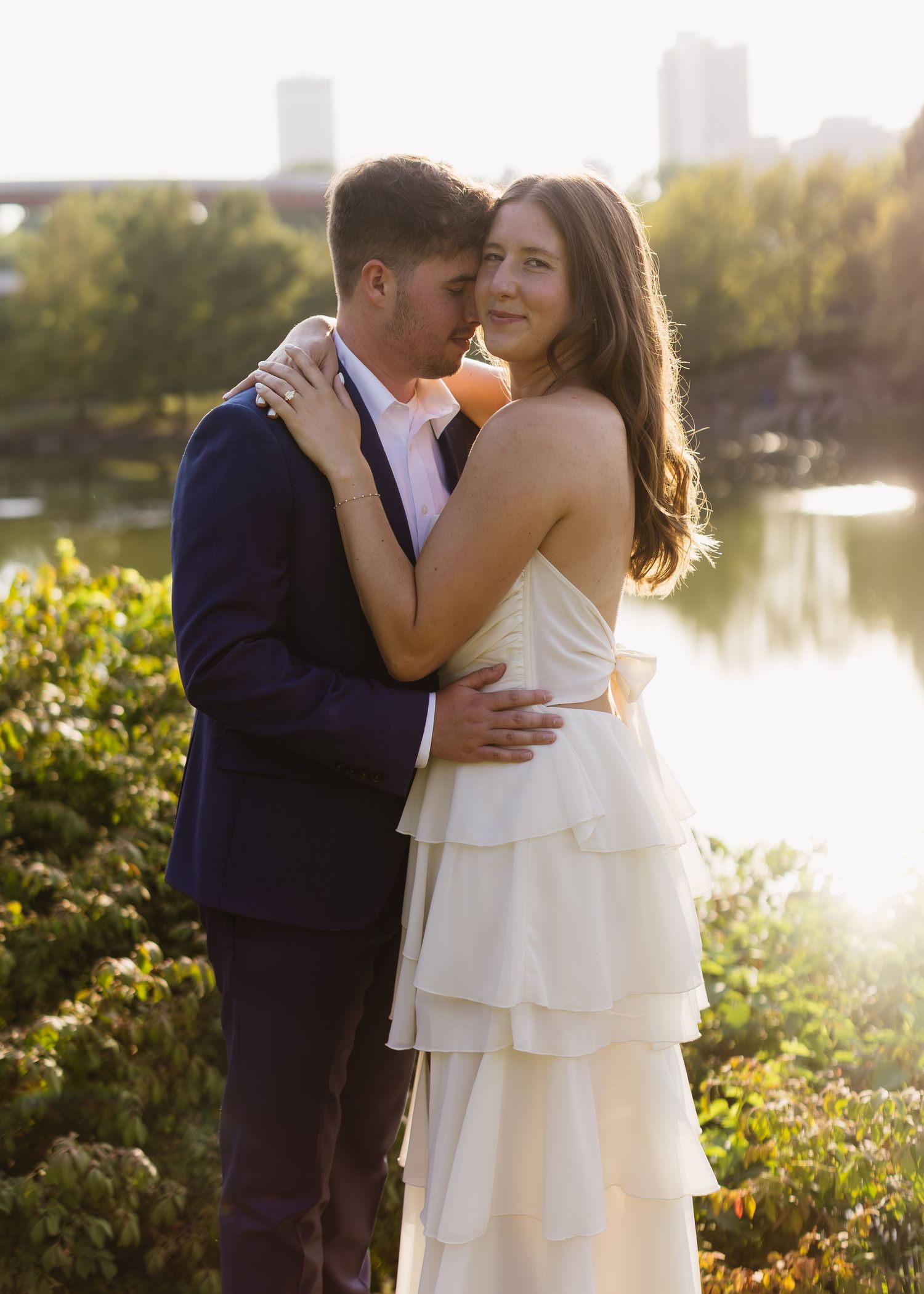 A romantic moment captured at sunset near a lake with city buildings in the distance.