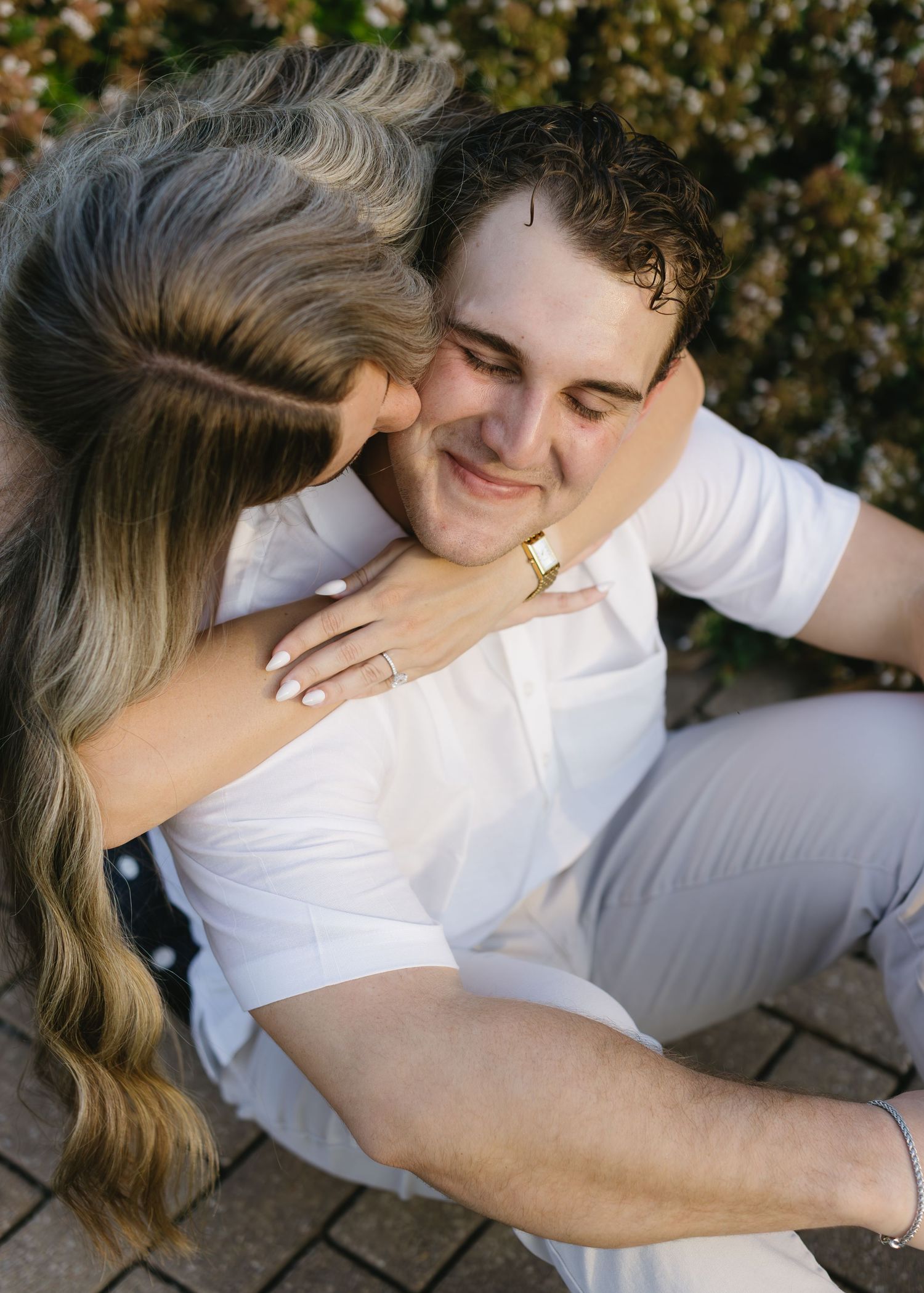 A couple shares an intimate moment outdoors with the woman kissing the man's cheek as he smiles in a white shirt.