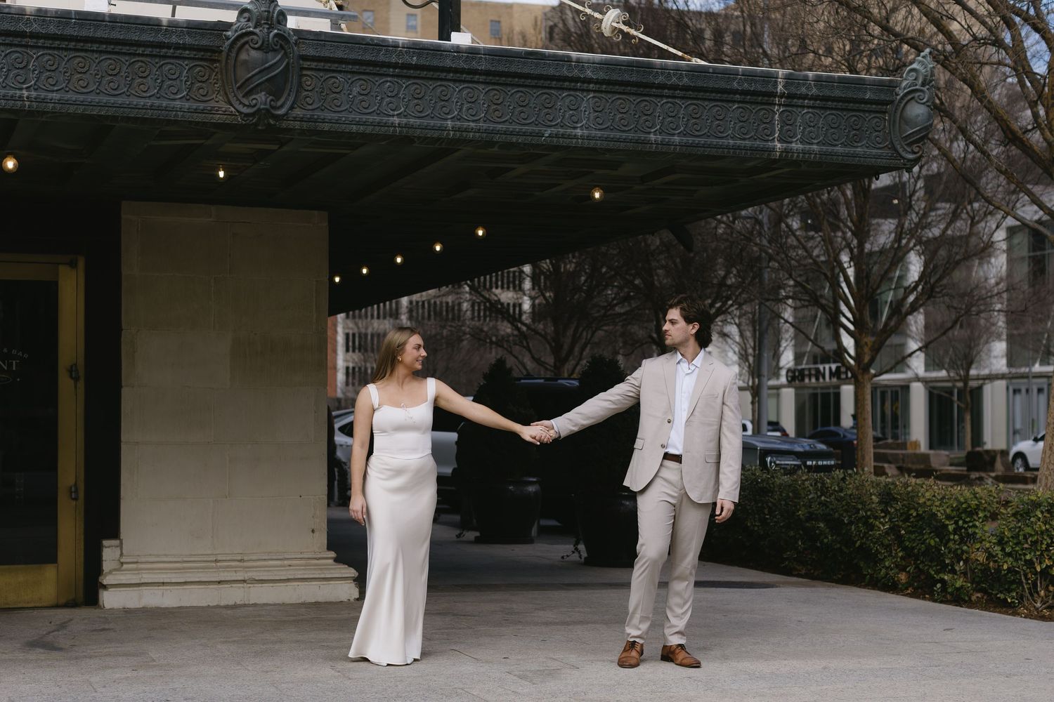 A couple in beige and white attire holds hands while walking under string lights outdoors.