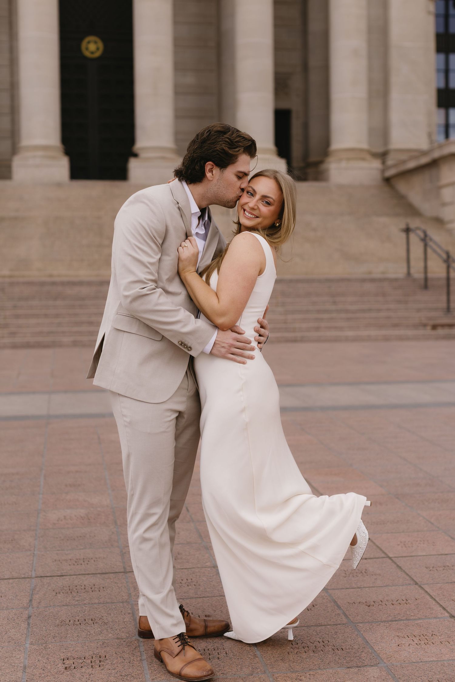 A romantic couple in wedding attire embrace and dance on stone steps outside a grand building.