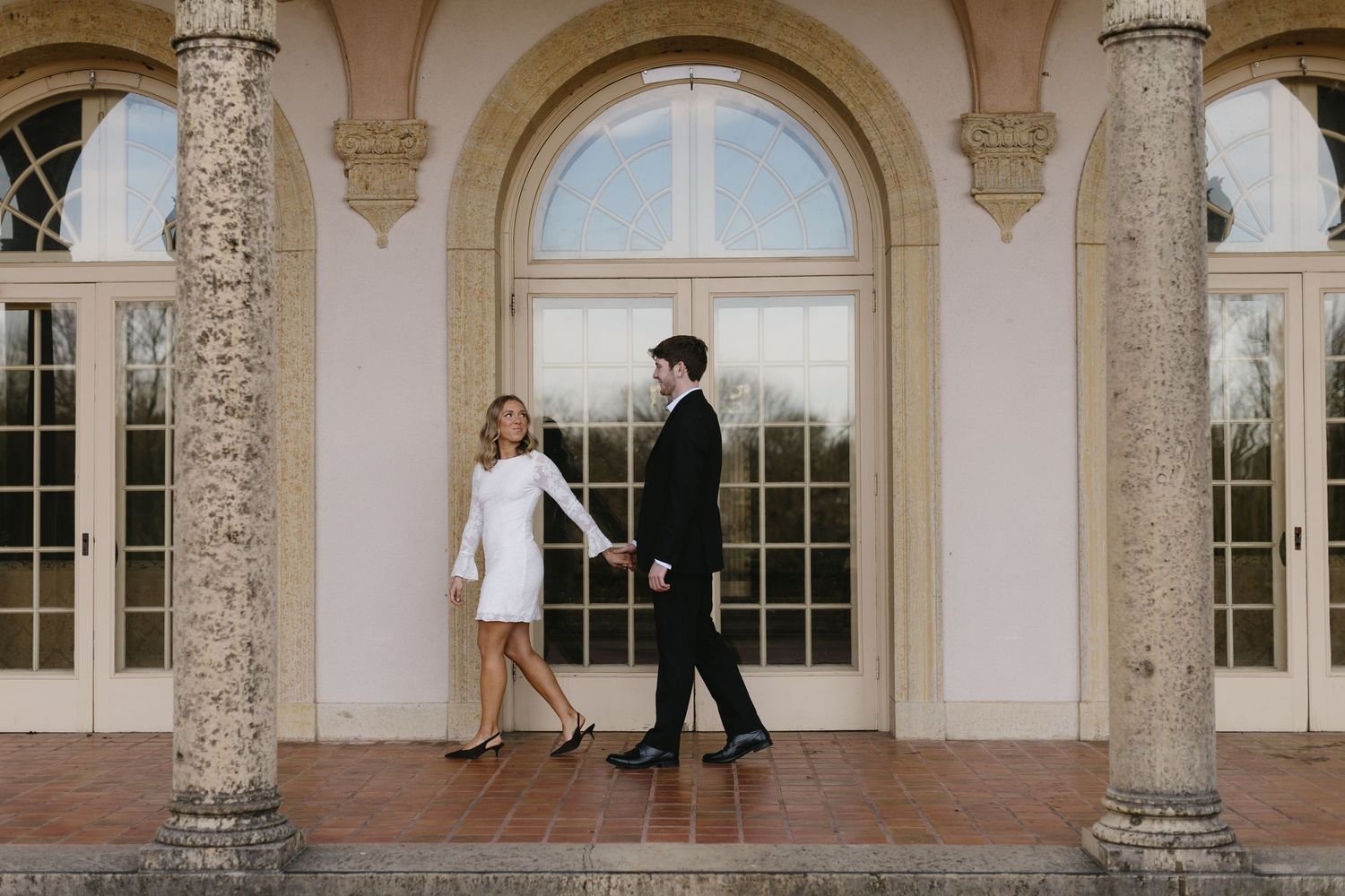 Elegant wedding couple stands between ornate stone columns at a grand estate venue during sunset.