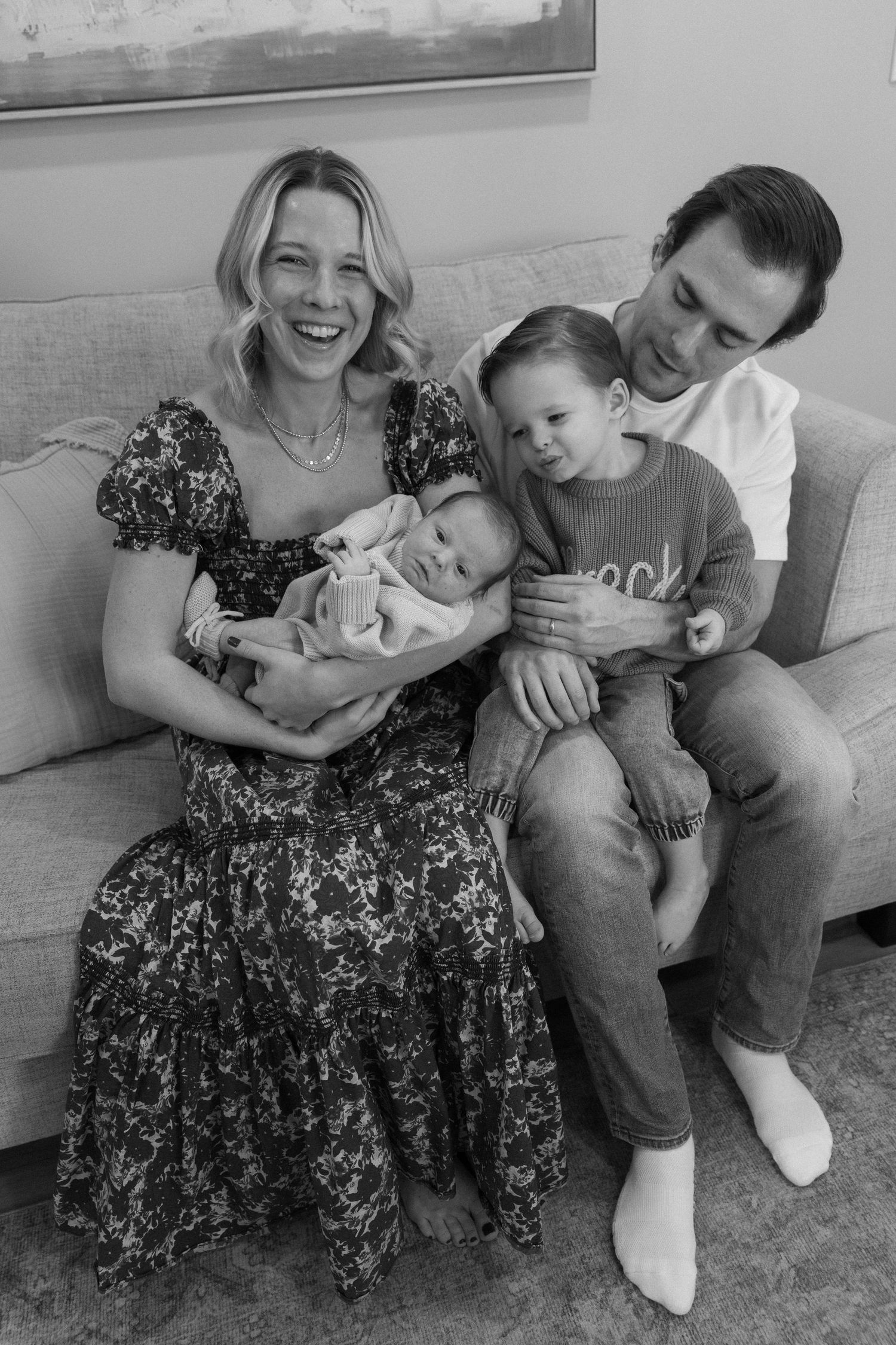 A couple sits together on a grey couch, sharing joyful moments with their newborn baby.
