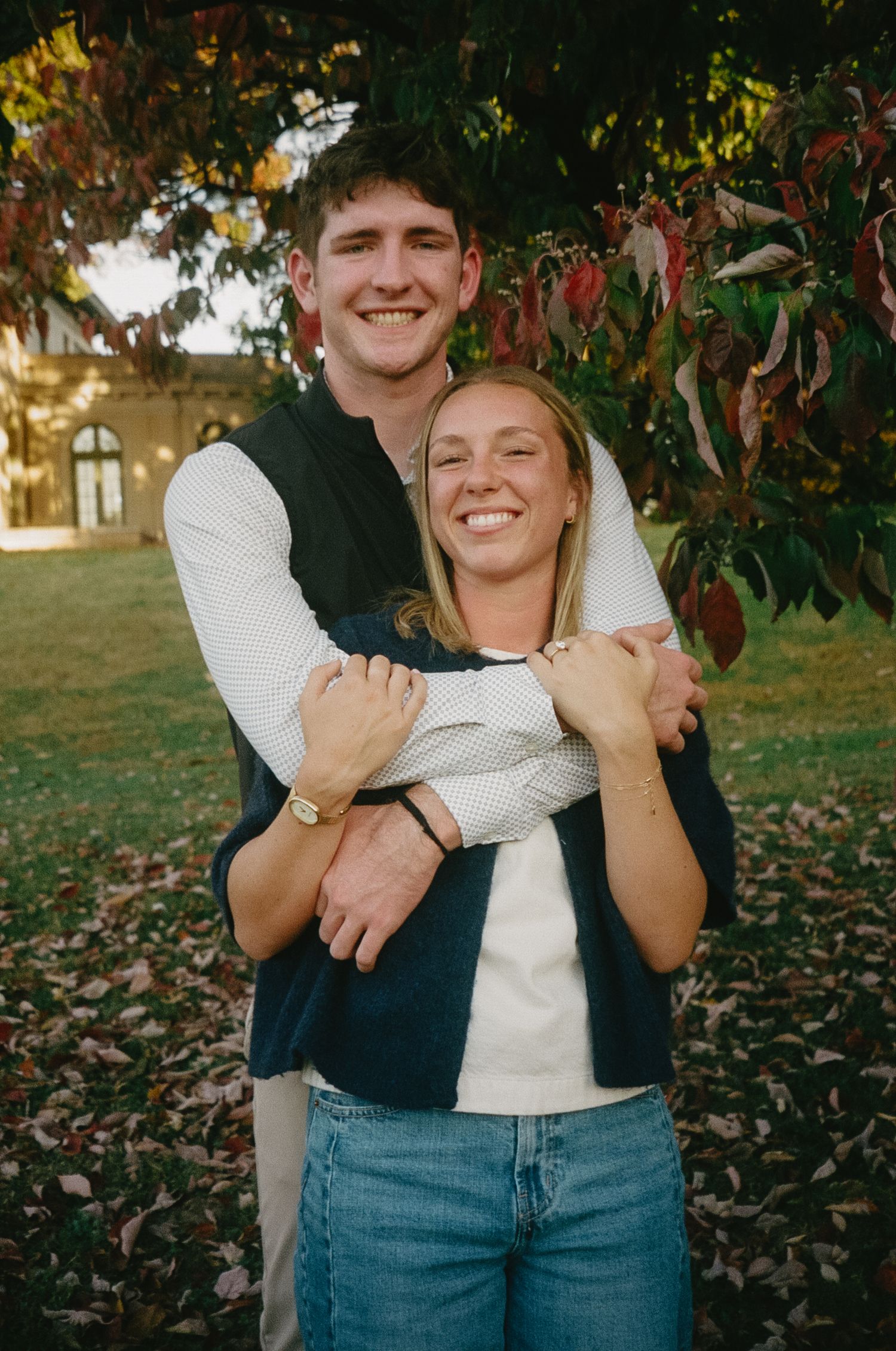 A couple in casual attire embraces playfully in a fall setting with autumn leaves on the ground.