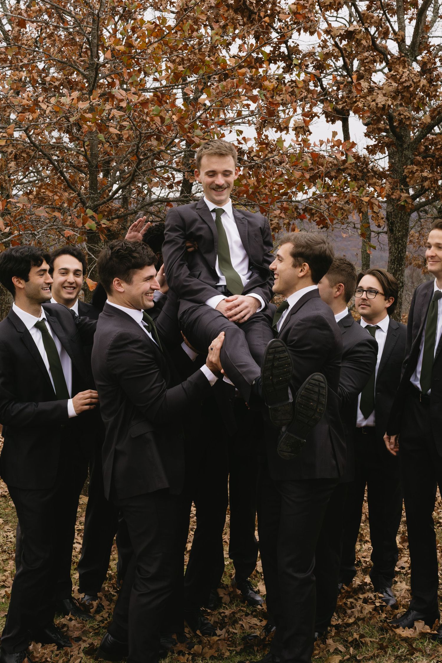 Groomsmen in black suits and ties gather for a group photo outdoors among fall foliage.