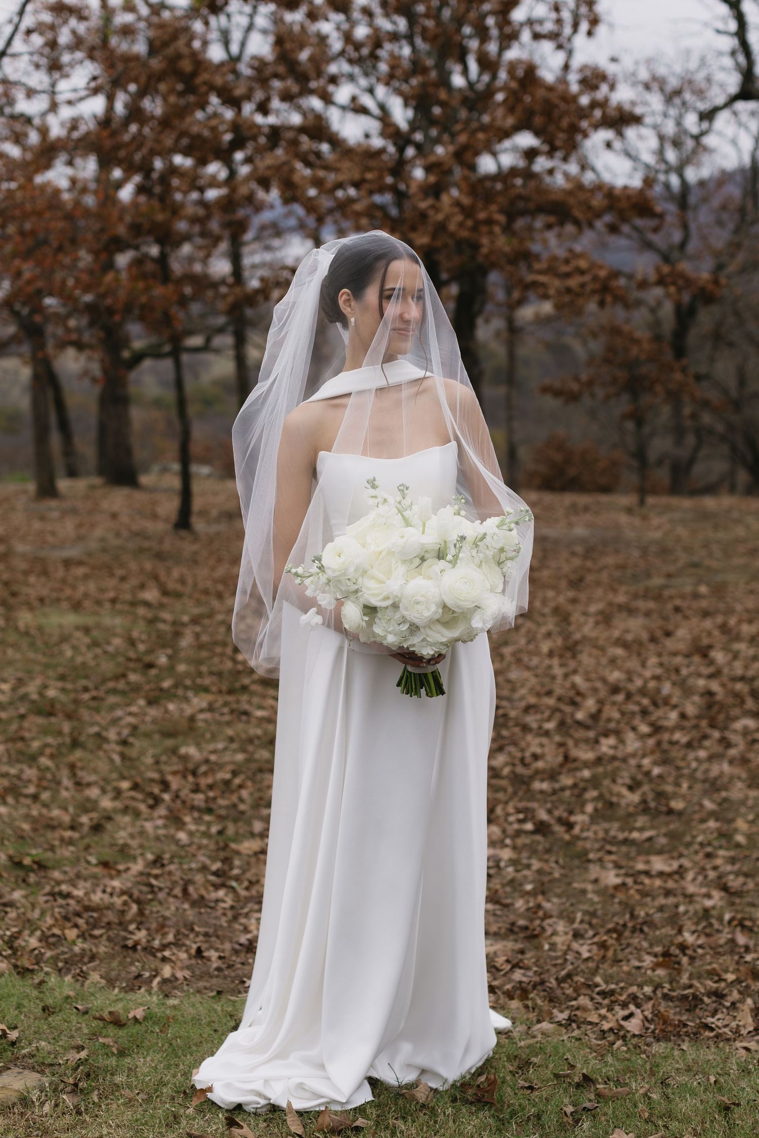 White wedding dress and veil silhouetted against autumn trees and fallen leaves.