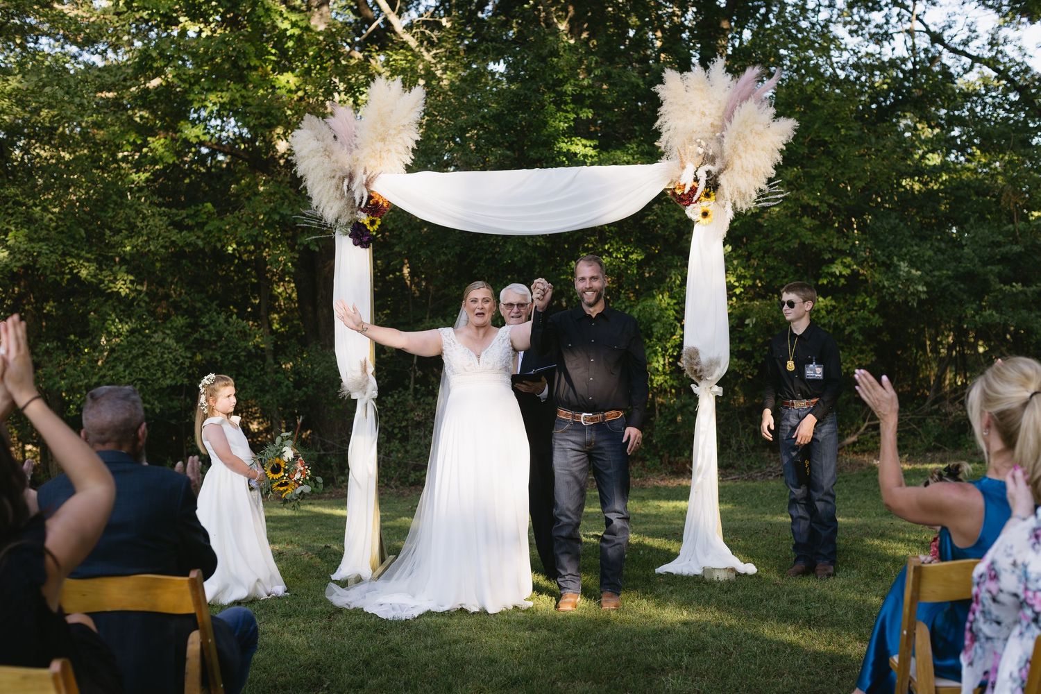 Outdoor wedding ceremony setup with white drapes and chairs under tall trees.