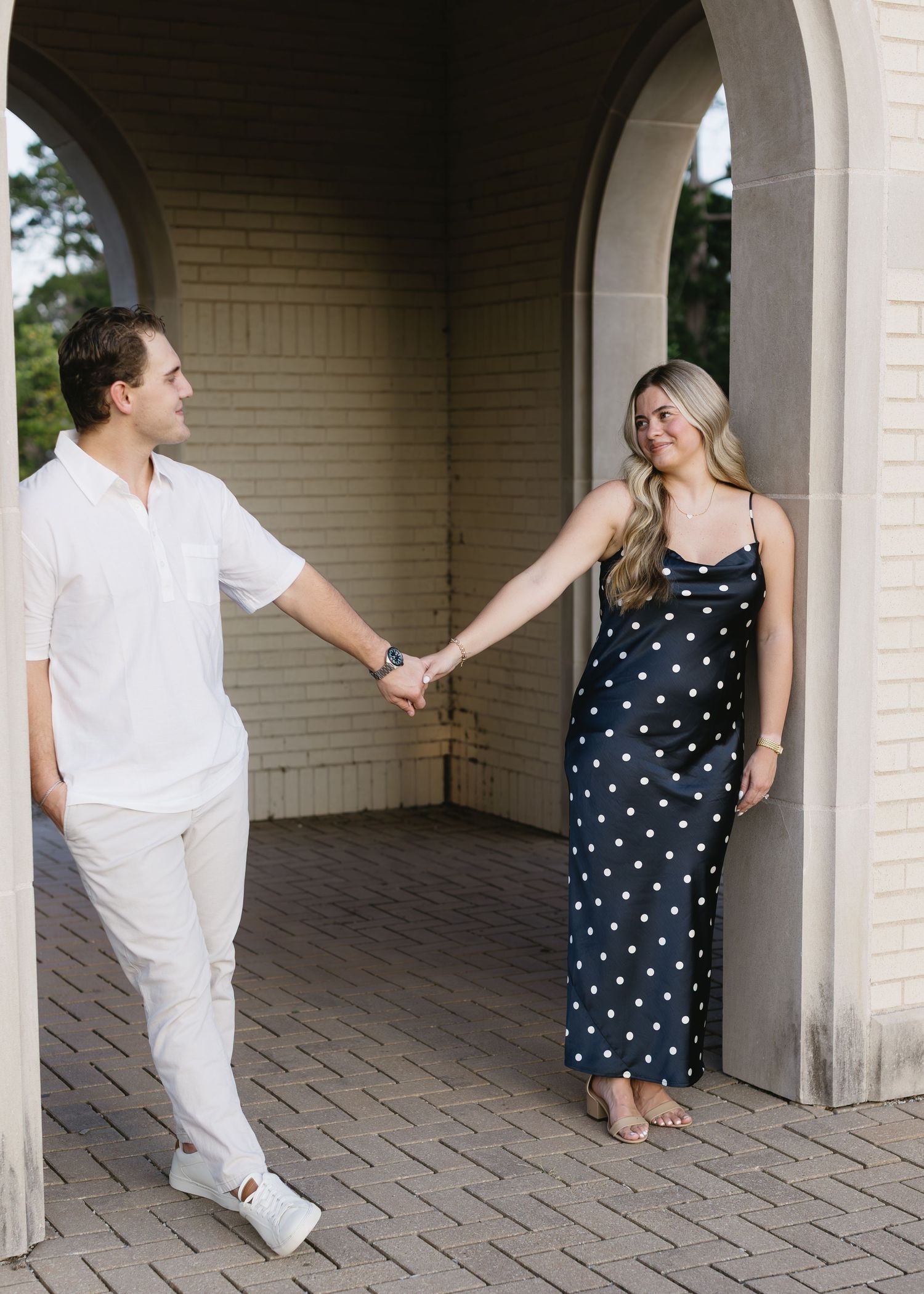 A couple holds hands under an archway, with one wearing white and the other in a polka dot dress.