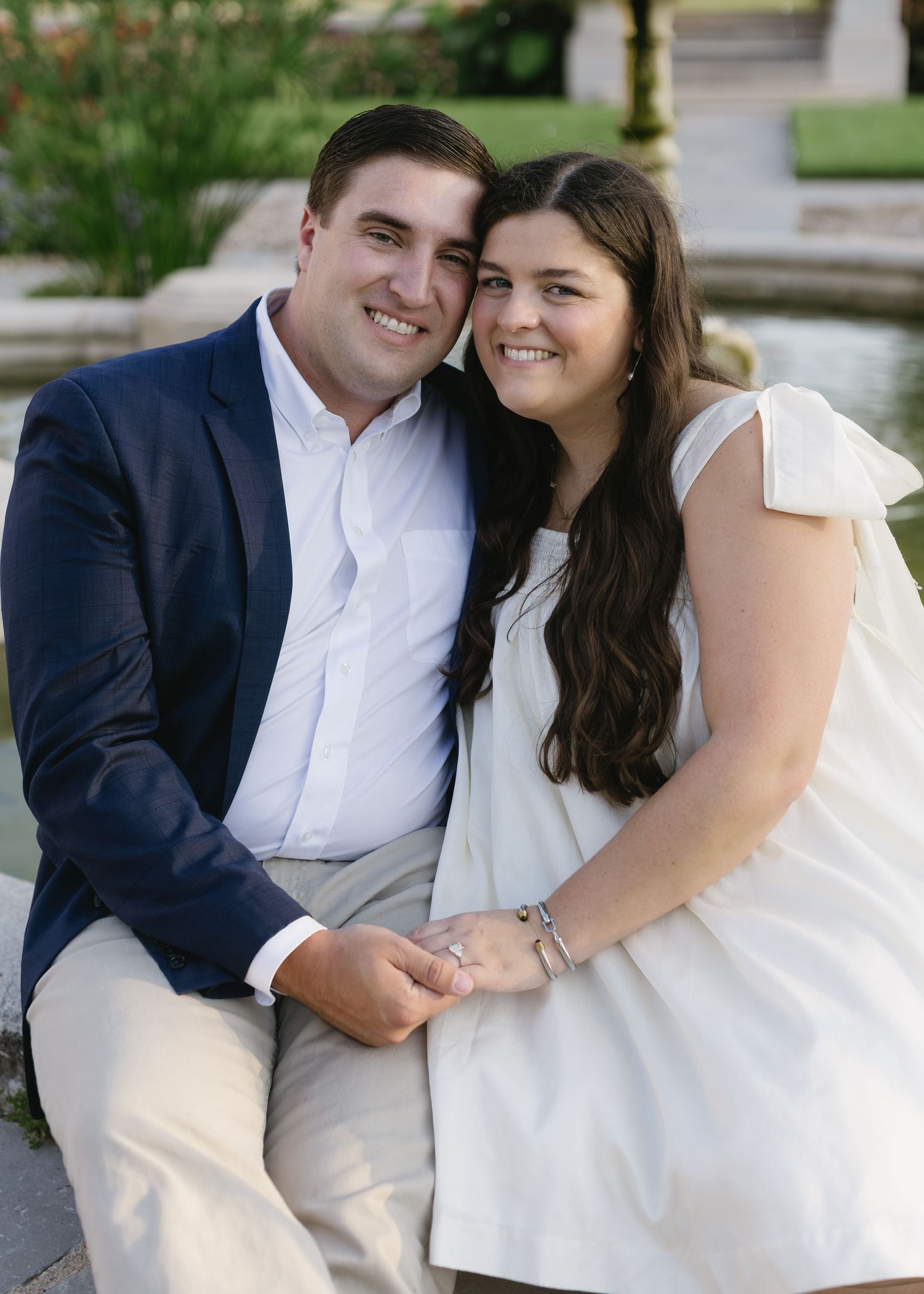 A couple in formal attire sitting outdoors by water, with the bride wearing a white dress and groom in navy suit.