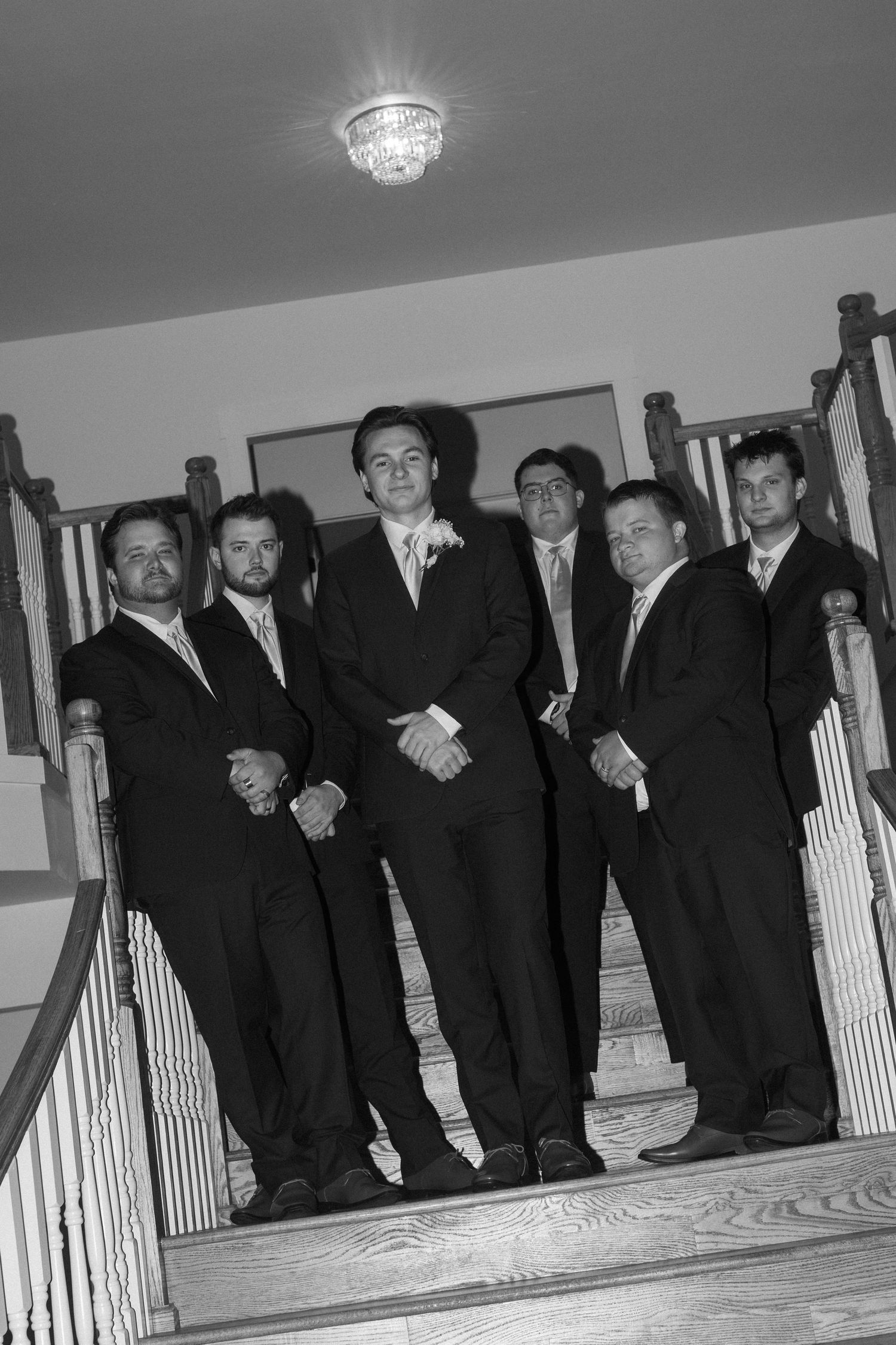 Groomsmen lined up on wooden stairs wearing dark suits in a fun candid black and white photo.