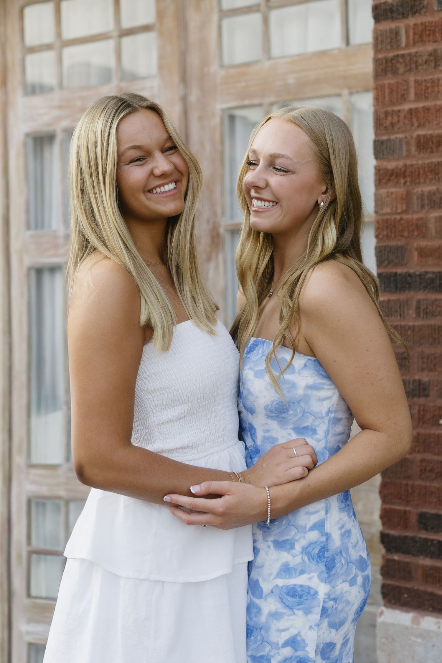 Two friends in white and blue dresses share joyful laughter against a brick wall.