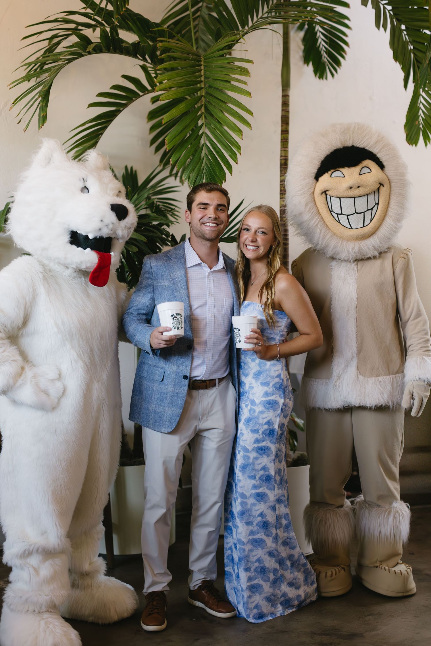 A couple poses with a large mascot character in white fluffy costume against a backdrop with tropical leaves.