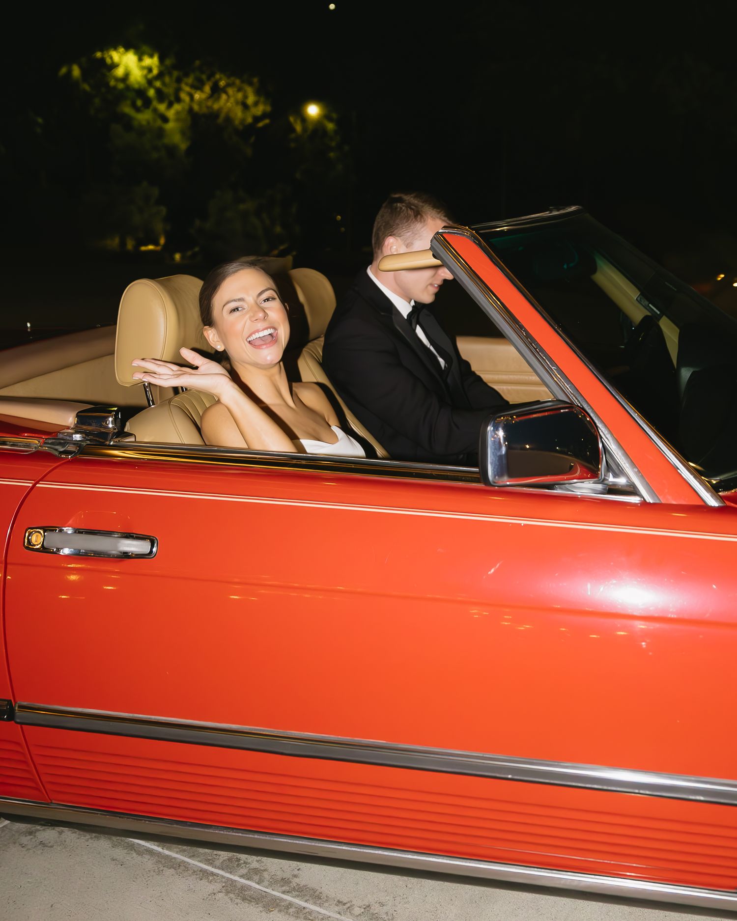 Newlyweds sharing a laugh while riding in a classic red convertible at night.