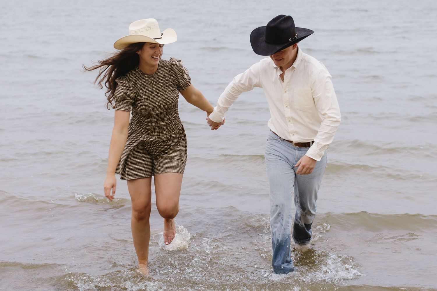 Playful couple splashes in ocean water while wearing western-inspired outfits.