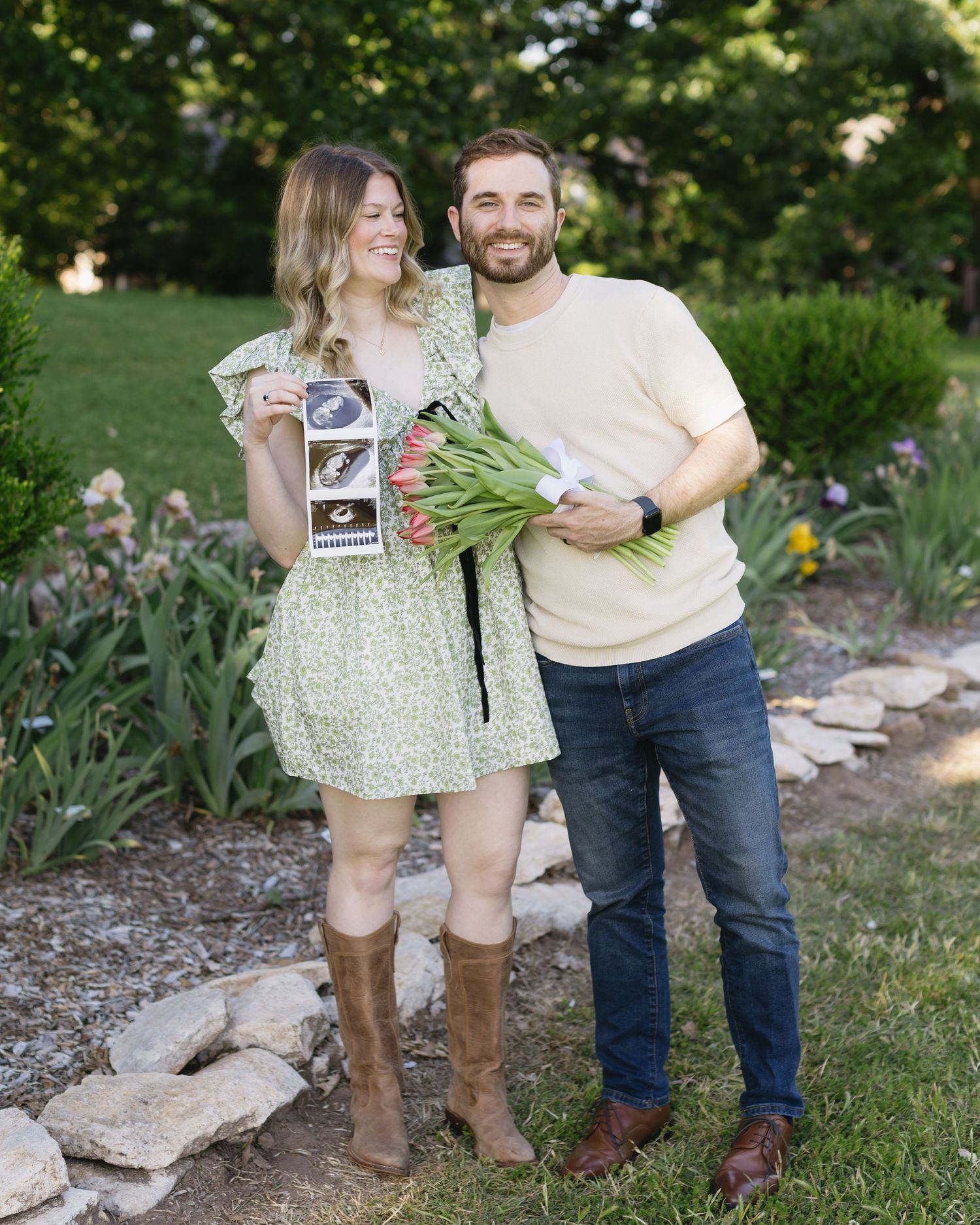 Couple shares pregnancy announcement photos while standing on garden path with flowers.
