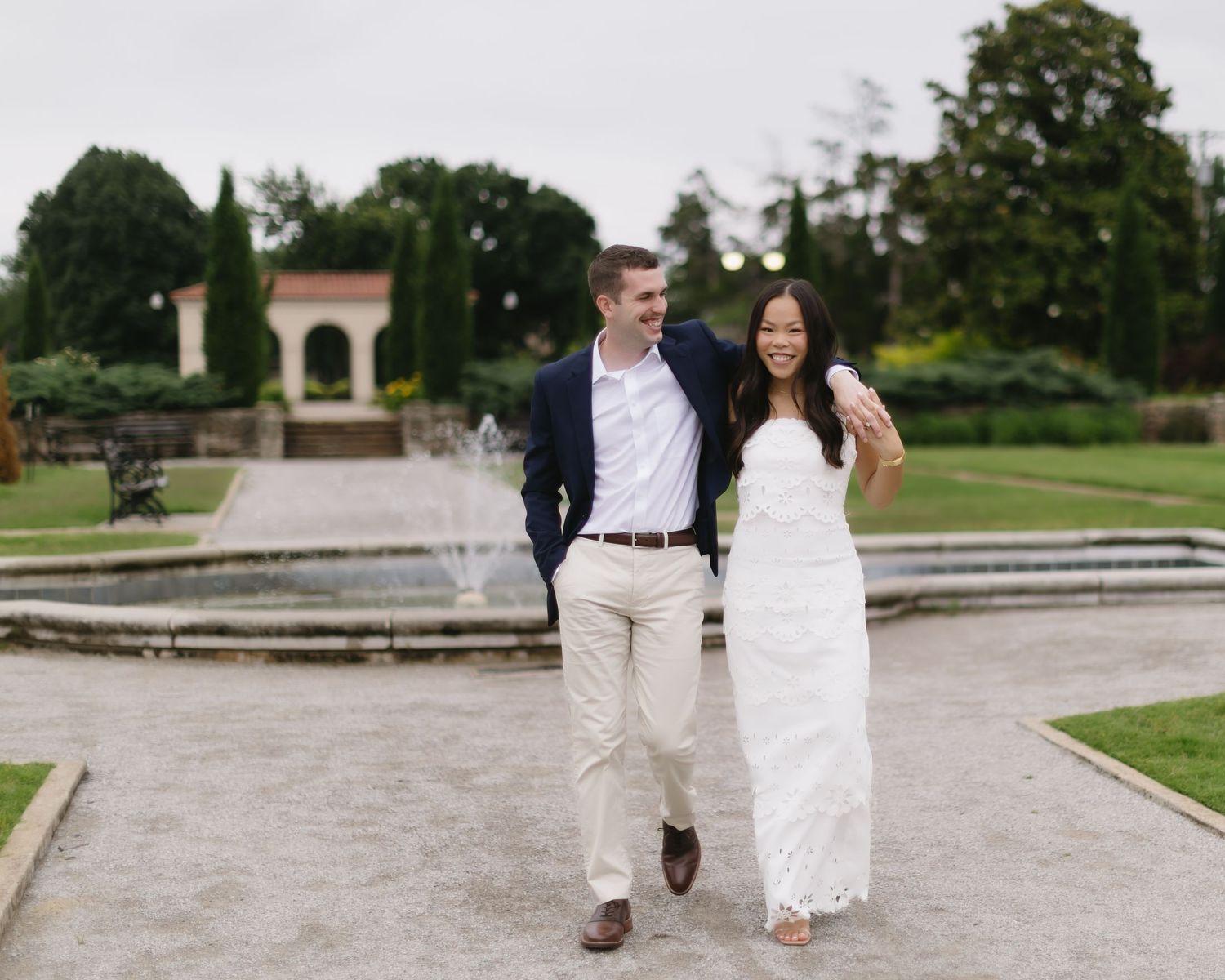 A couple in coordinated white and beige outfits strolls together on stone pavers in a formal garden setting.
