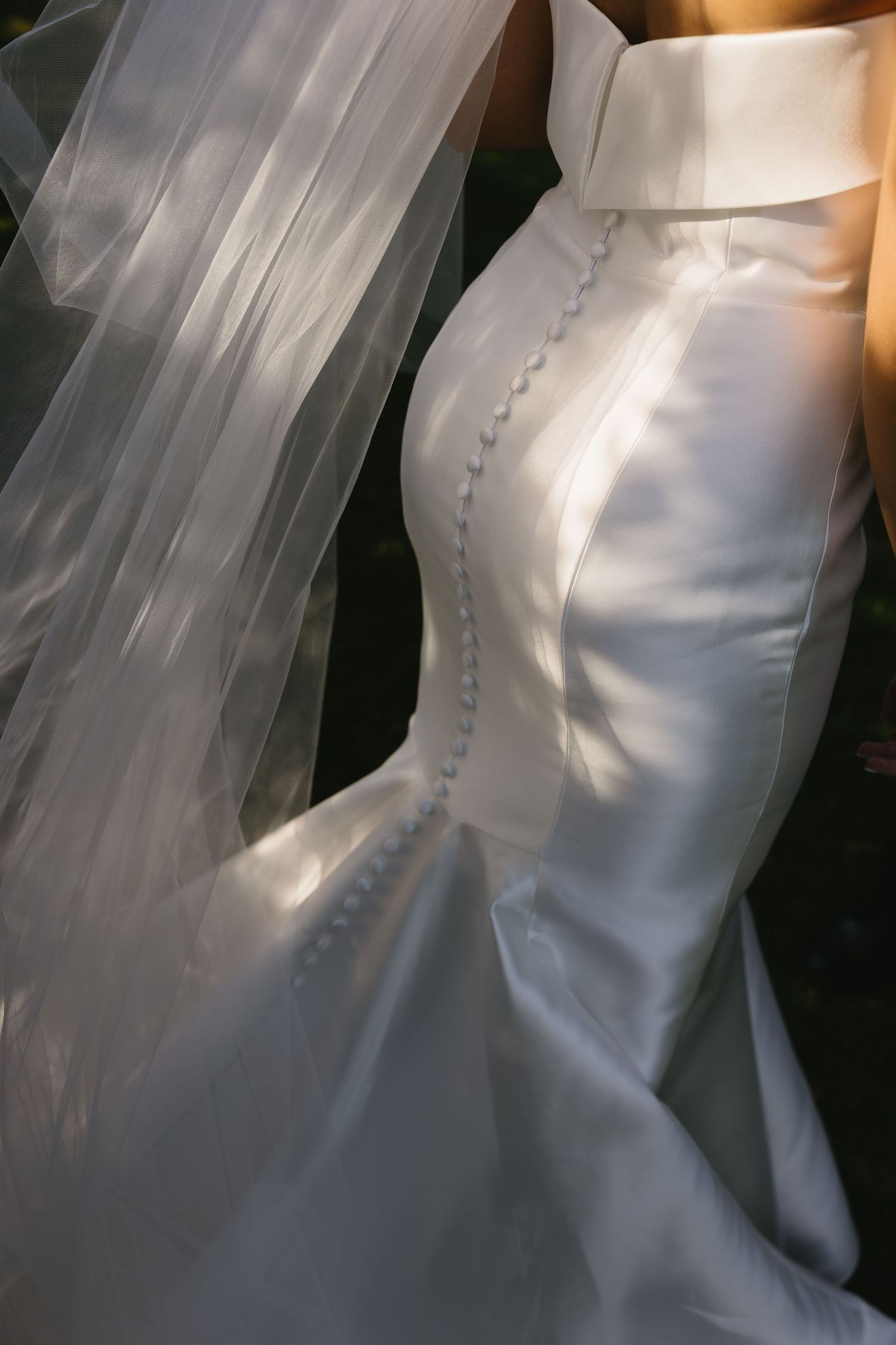 Close-up of a white satin wedding dress with decorative button details and flowing veil in soft lighting.