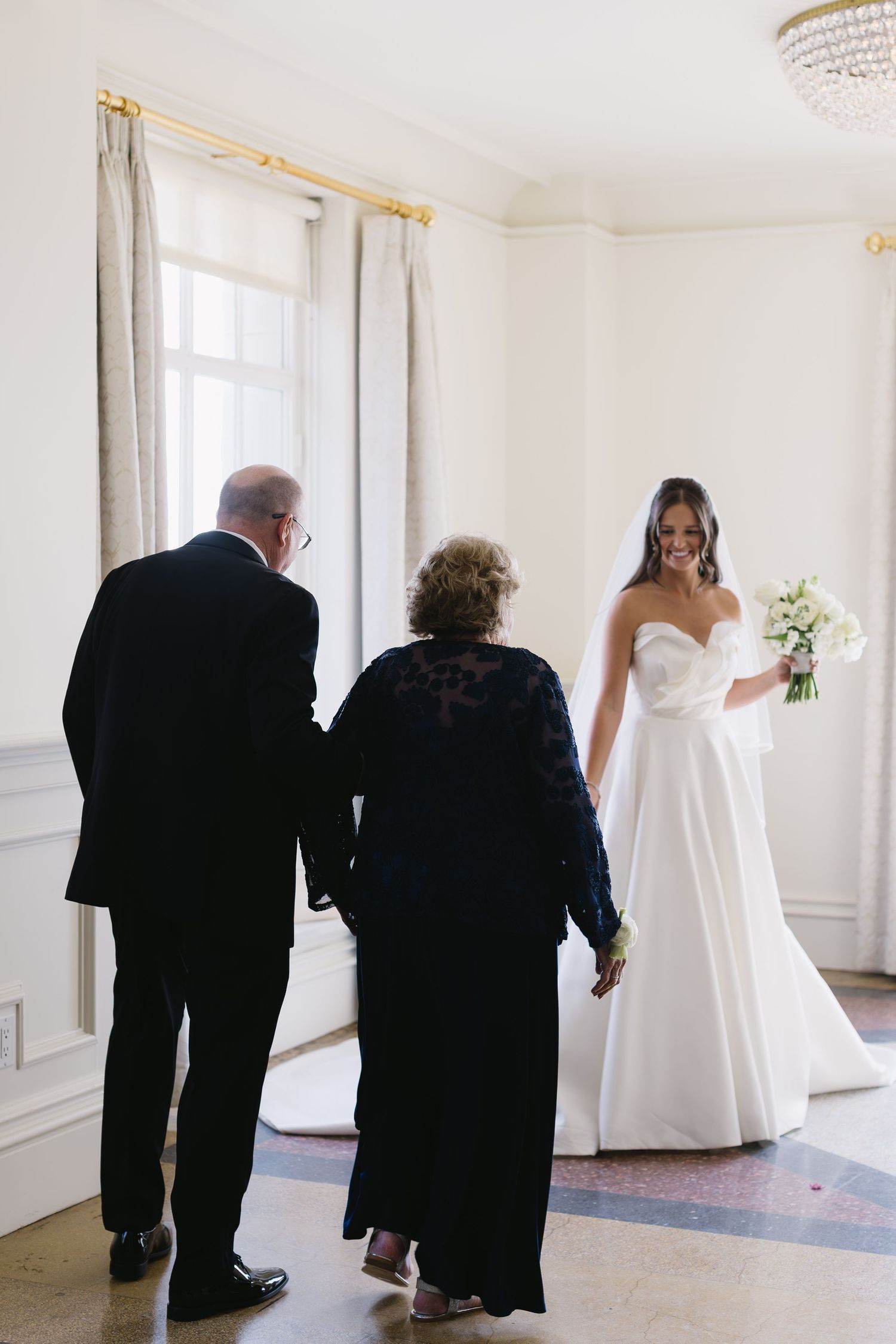 An elegant white wedding gown is showcased in a bright room with guests in formal black attire.