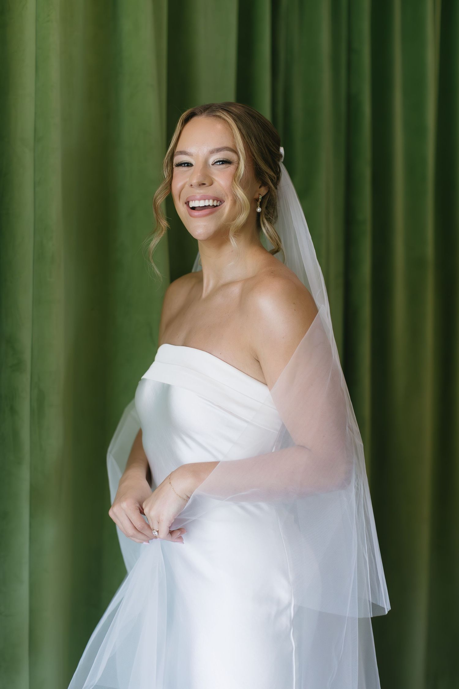 A bride in a classic white strapless wedding dress poses against an emerald green curtain backdrop.