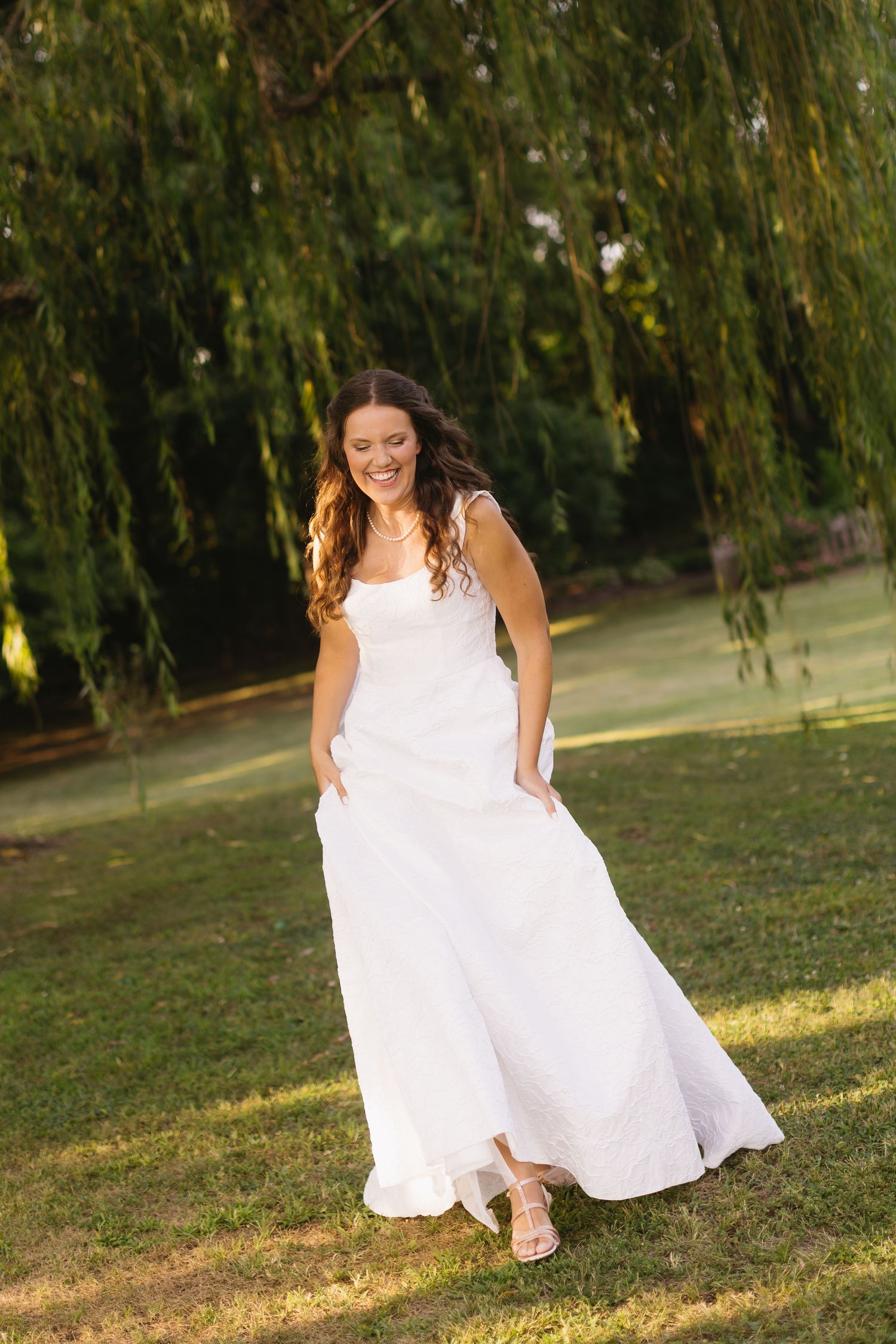 A bride twirls her white wedding dress in a sunlit garden with weeping willow trees.