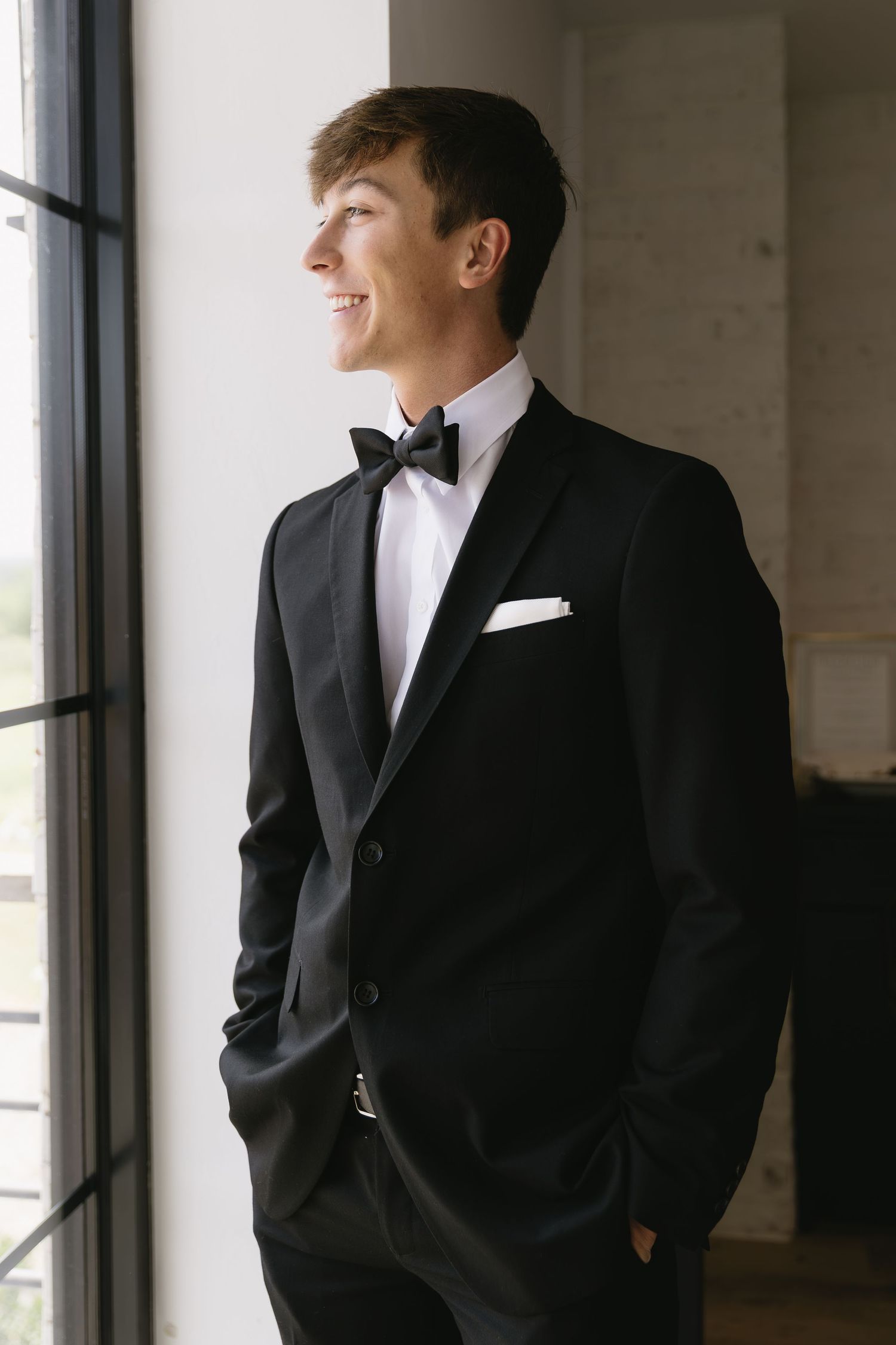Groomsmen in classic black tuxedos and bow ties pose by large windows.