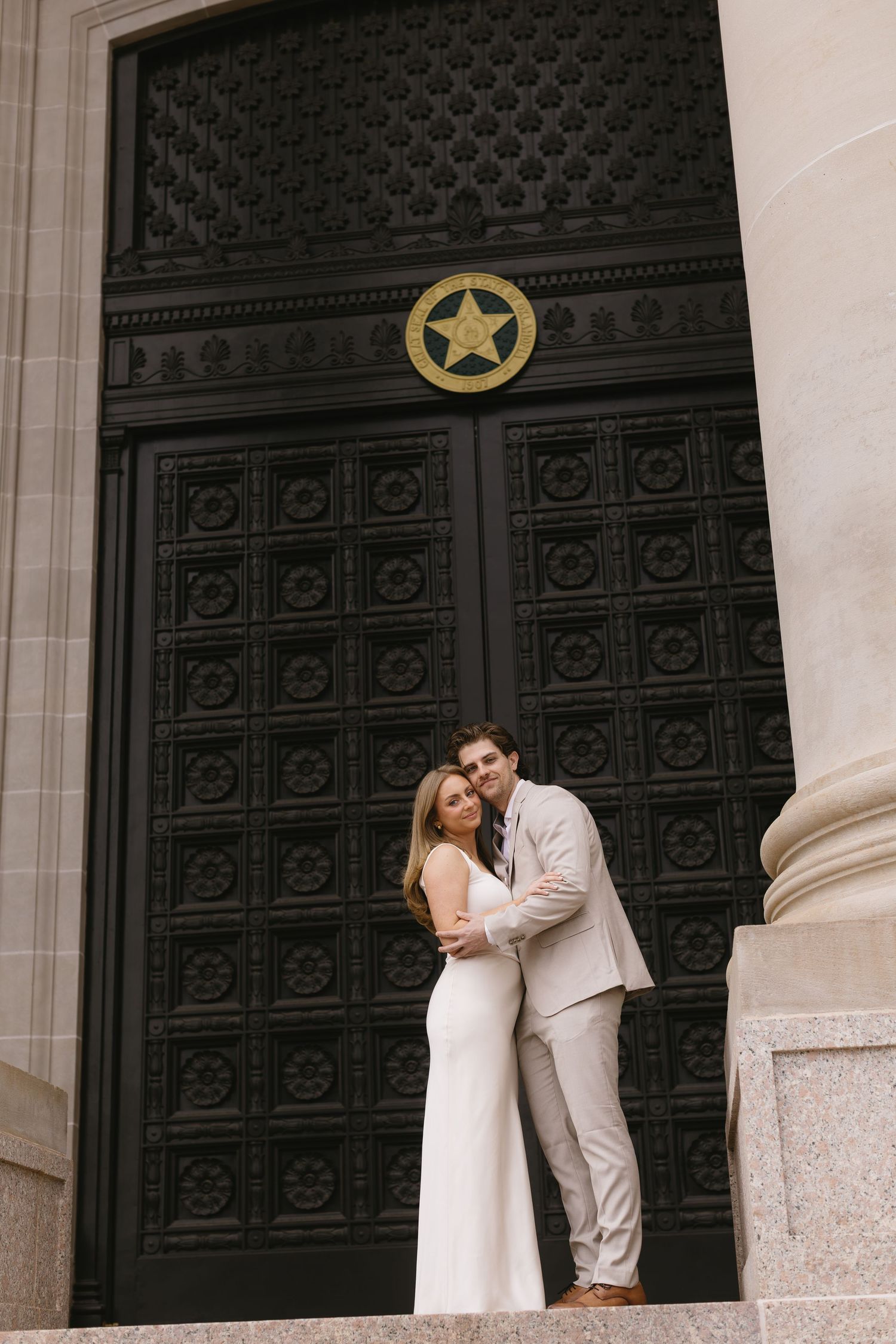 A couple poses romantically against ornate black doors and white columns of a historic building.