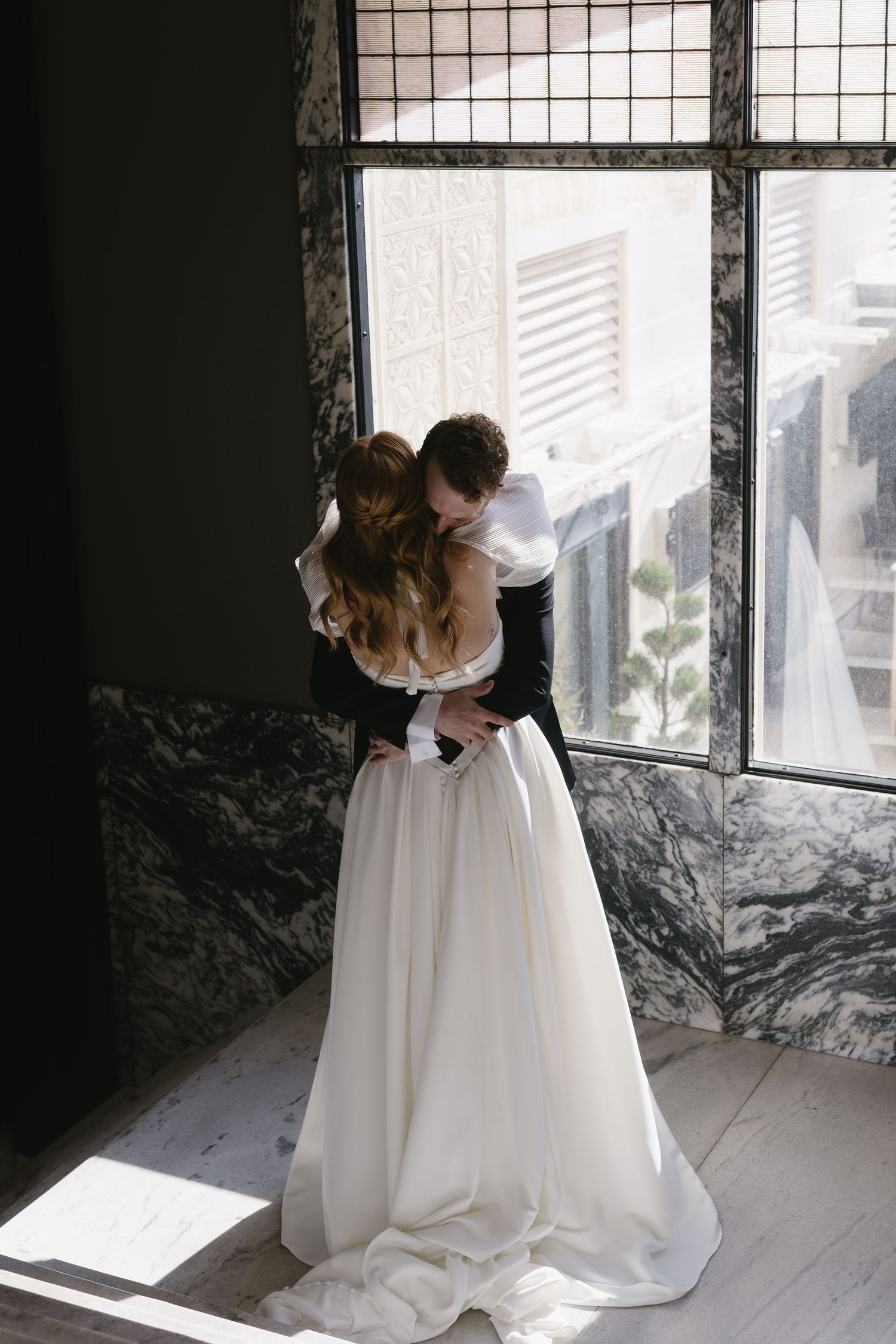Newlyweds share a tender embrace by large windows in modern architectural setting.